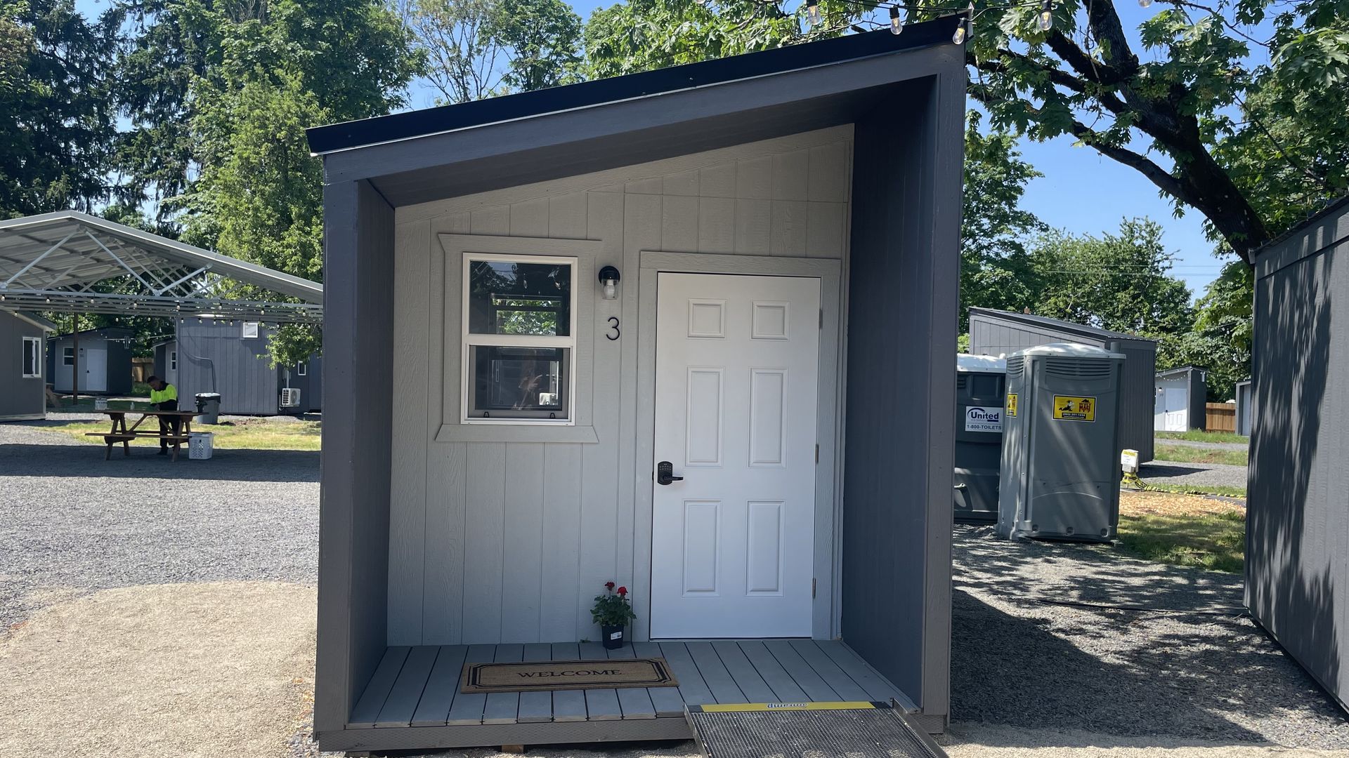 Small gray cabin numbered 3 with white door and window, a welcome mat, and a potted plant on the porch; gravel area and portable toilets visible in background under sunny blue sky.