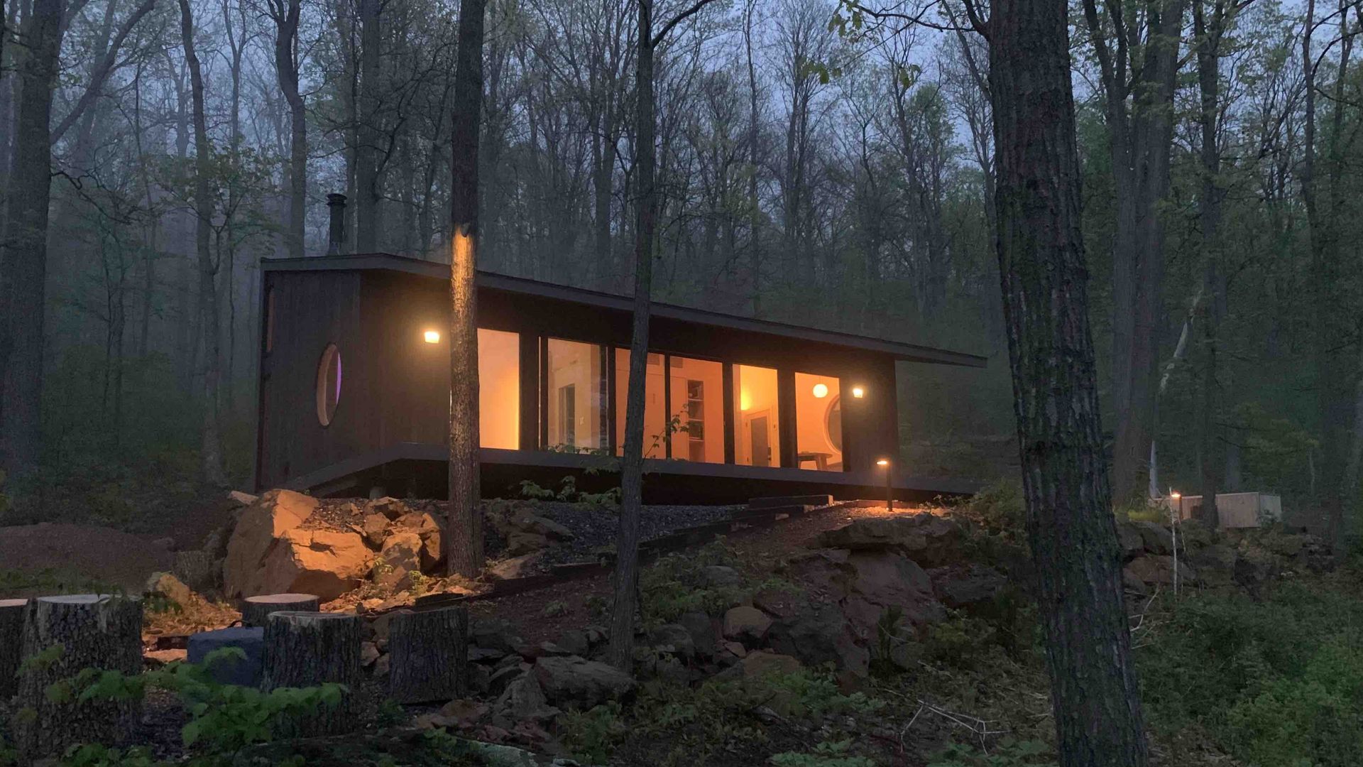 Modern dark cabin with glass walls sits on a rocky hillside inside a misty forest. Warm interior lights glow through large windows; surrounding trees and small exterior lamps line a path.