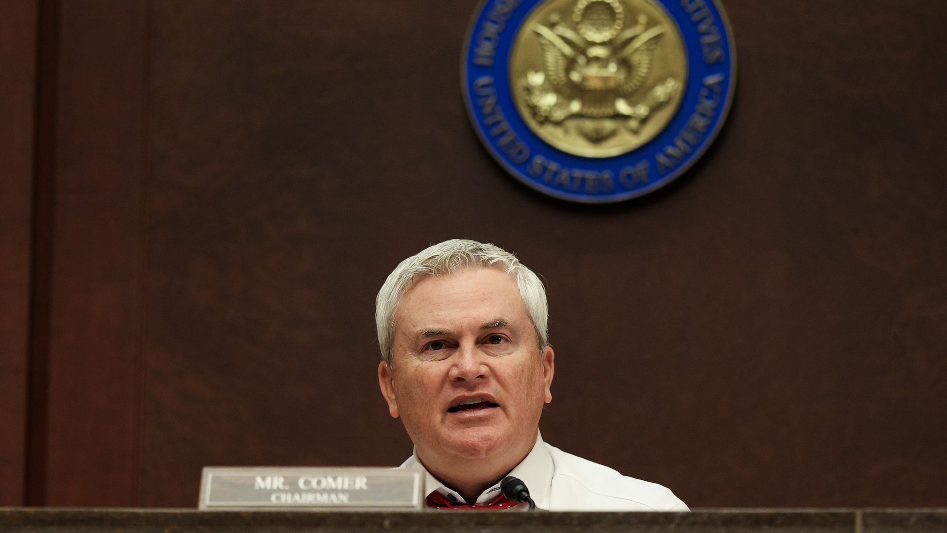 James Comer sitting in a brown wooden room with a gold and blue seal on it, wearing a white shirt.