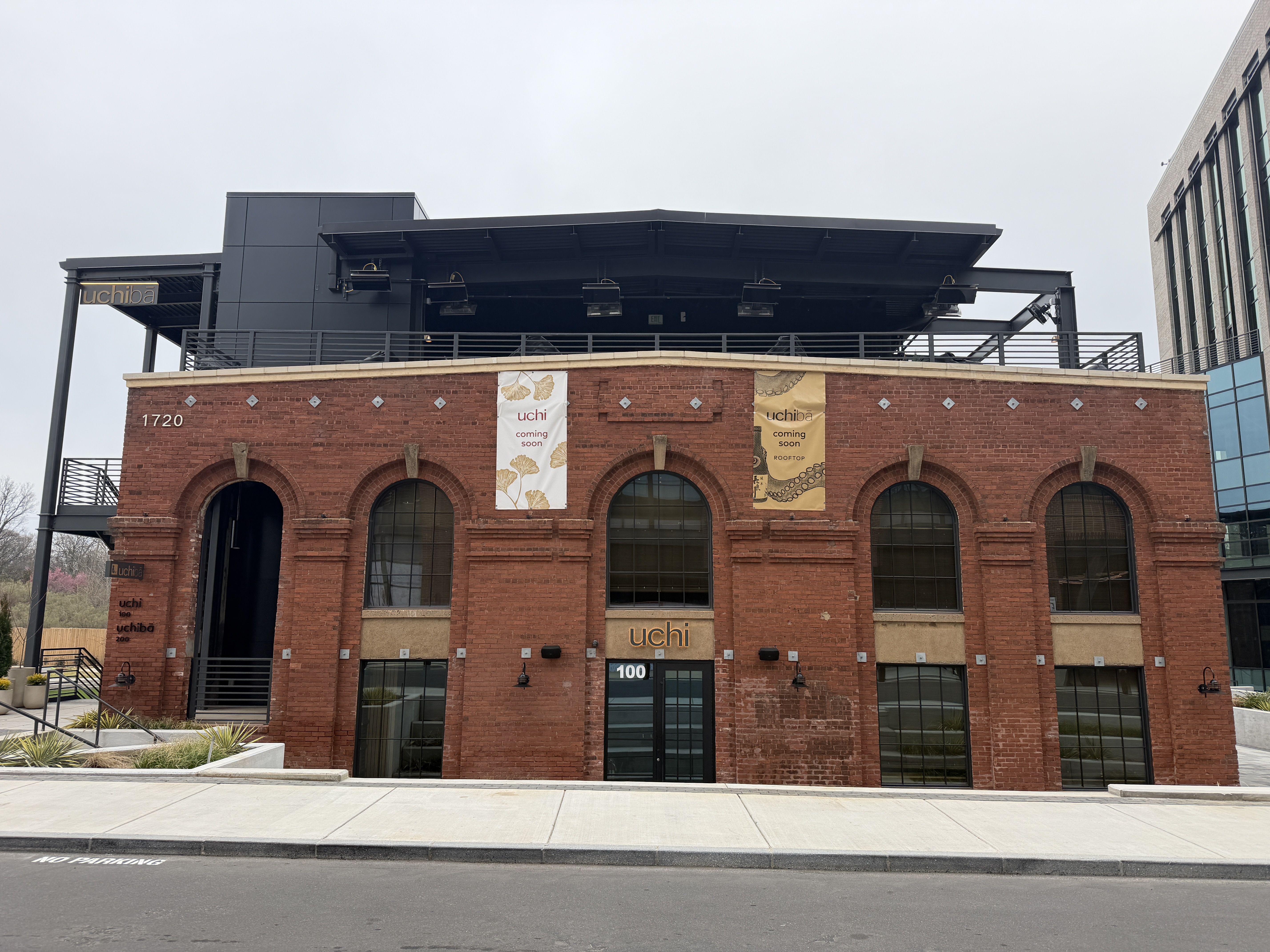 Red brick building with arched windows and black metal upper structure, two banners reading "uchi coming soon" and address numbers 1720 and 100 visible, overcast sky background.
