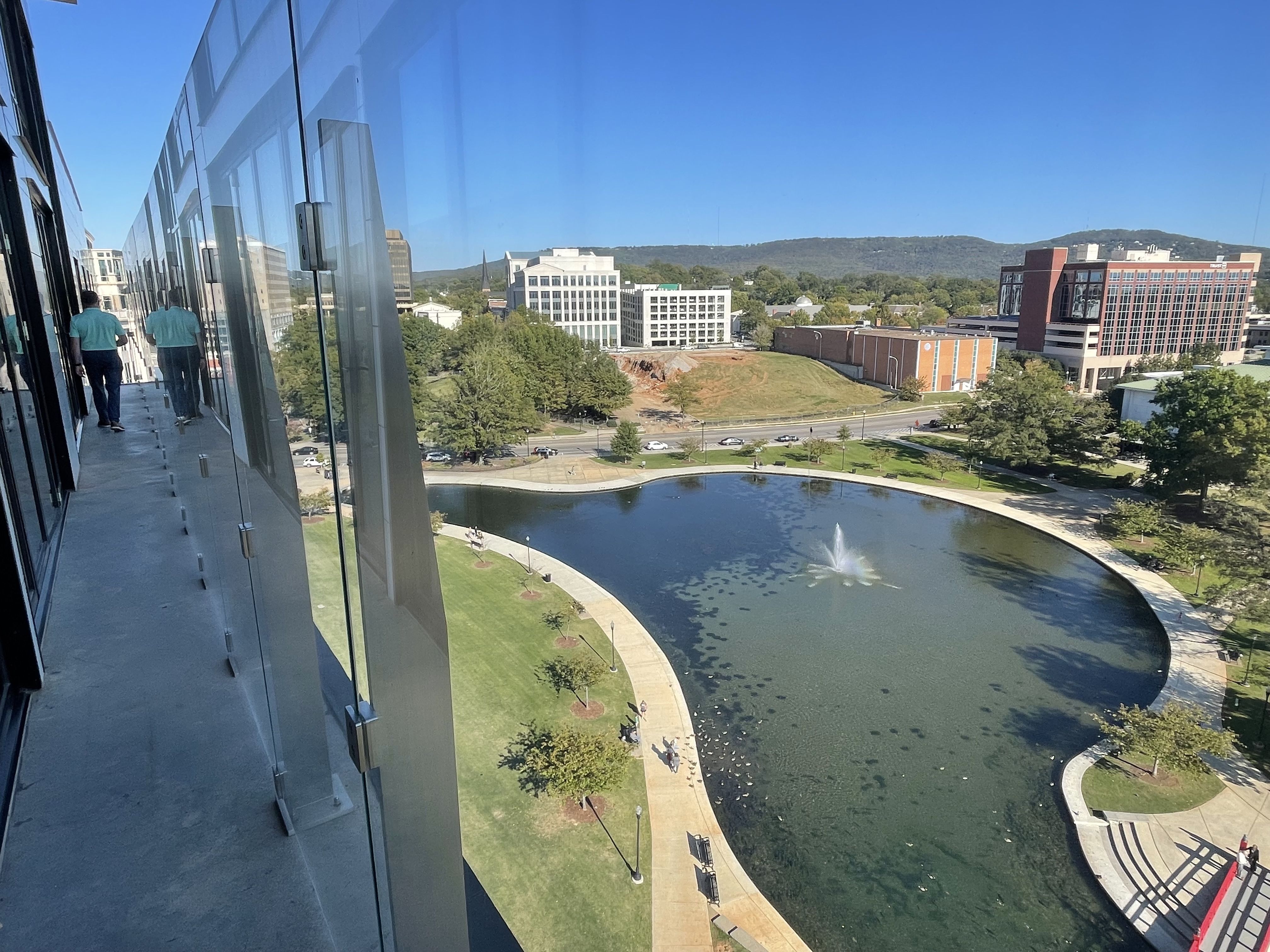 View from building balcony showing a man in a teal shirt reflected on glass, overlooking a park with a pond, fountain, pathways, benches, trees, cars on a nearby street, and hills under a clear blue sky.