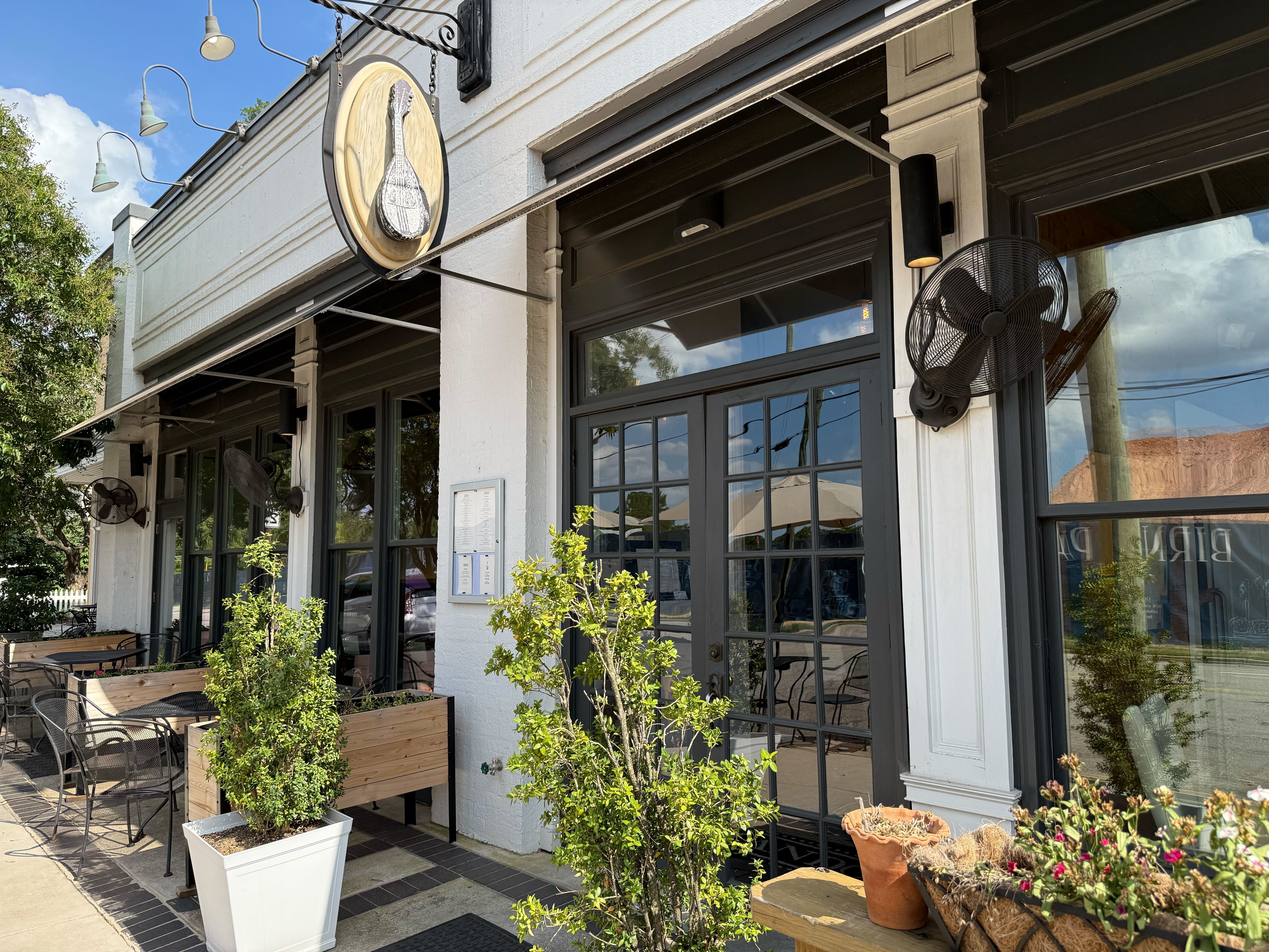 Outdoor seating area of a cafe with black metal chairs and tables, potted plants, and a sign featuring a stringed instrument on a white building under a blue sky.