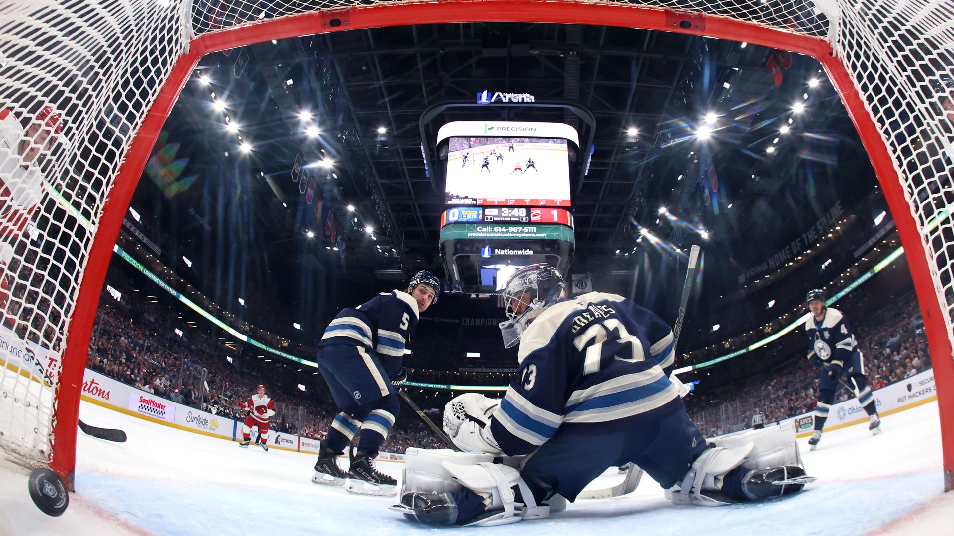 From behind the red goal frame, a Blue Jackets goalie blocks a shot as teammates and an opponent skate near the crease, with a packed arena and a large scoreboard overhead.