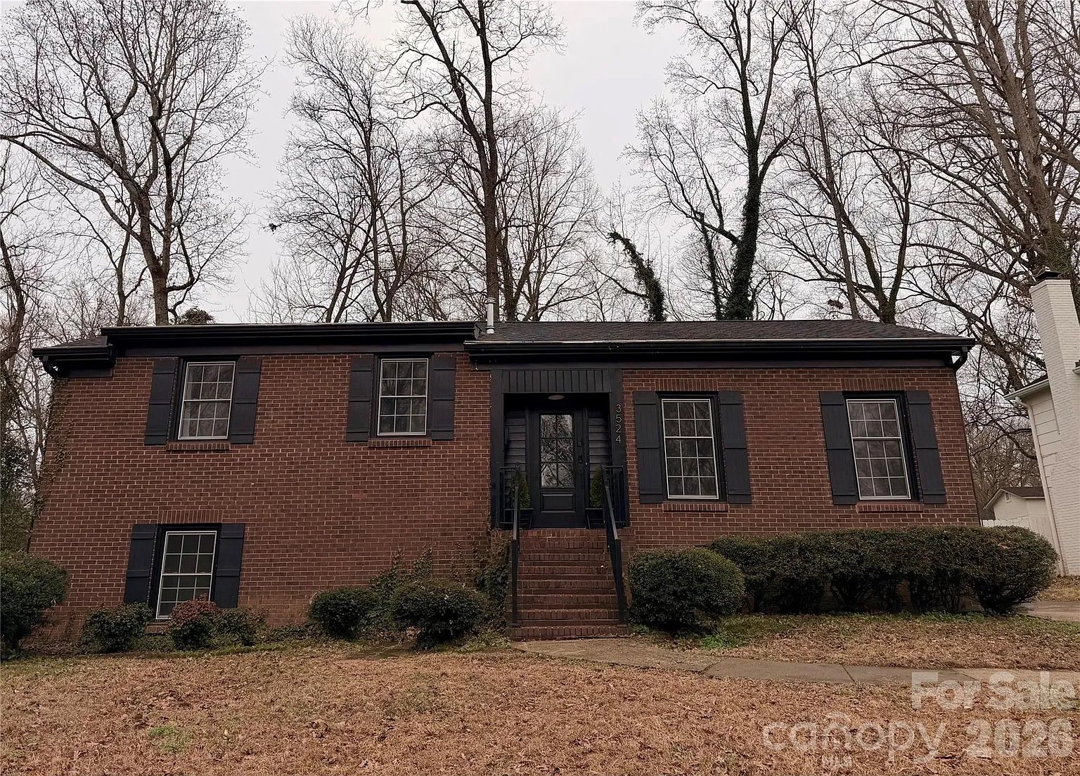 Single-story brick house with black shutters and a black front door, surrounded by leafless trees and shrubs, under overcast sky with dry grass in front.