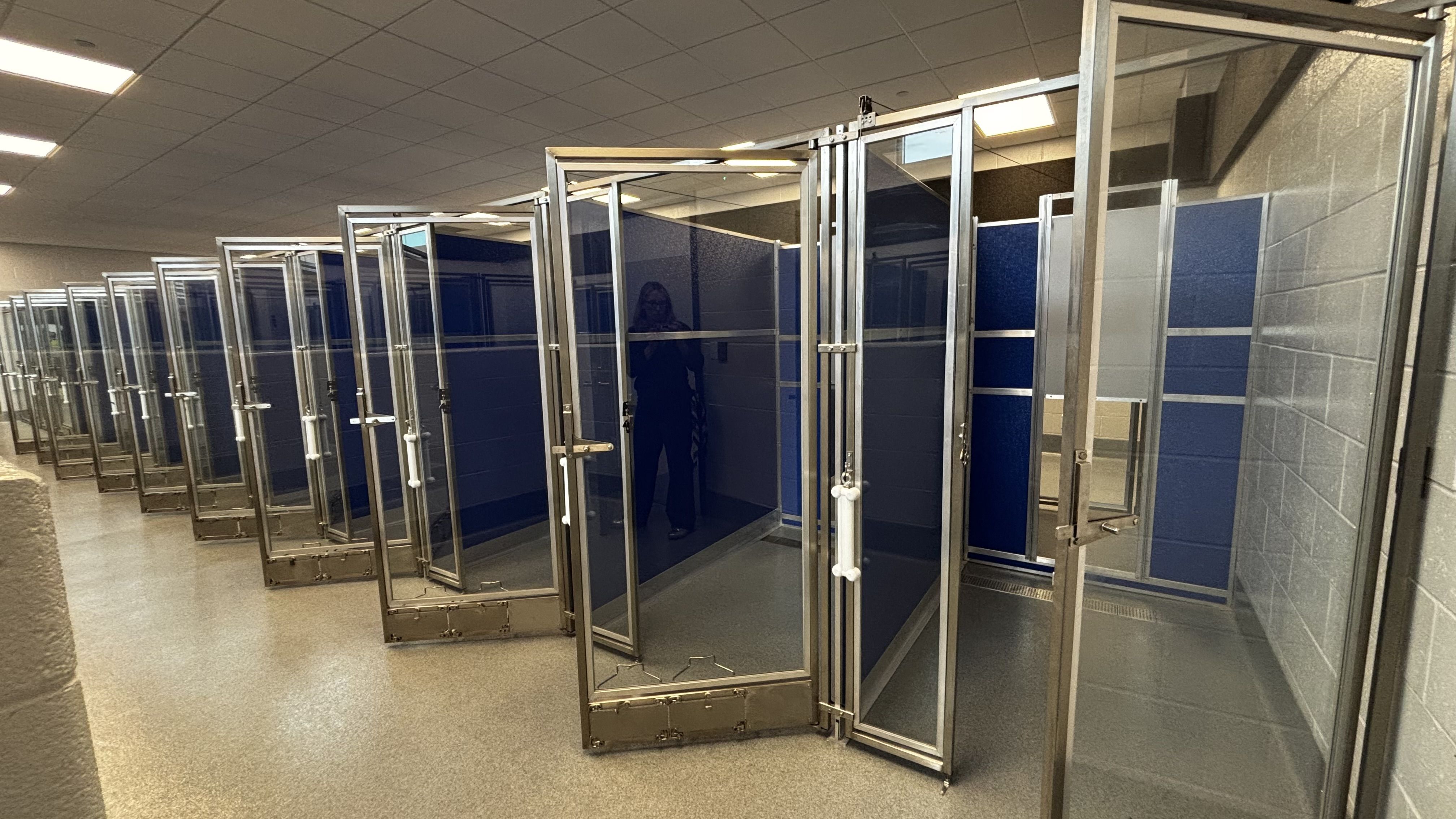 Long row of glass-and-metal jail cells with blue panels line a lit hallway; a person stands inside a distant cell.