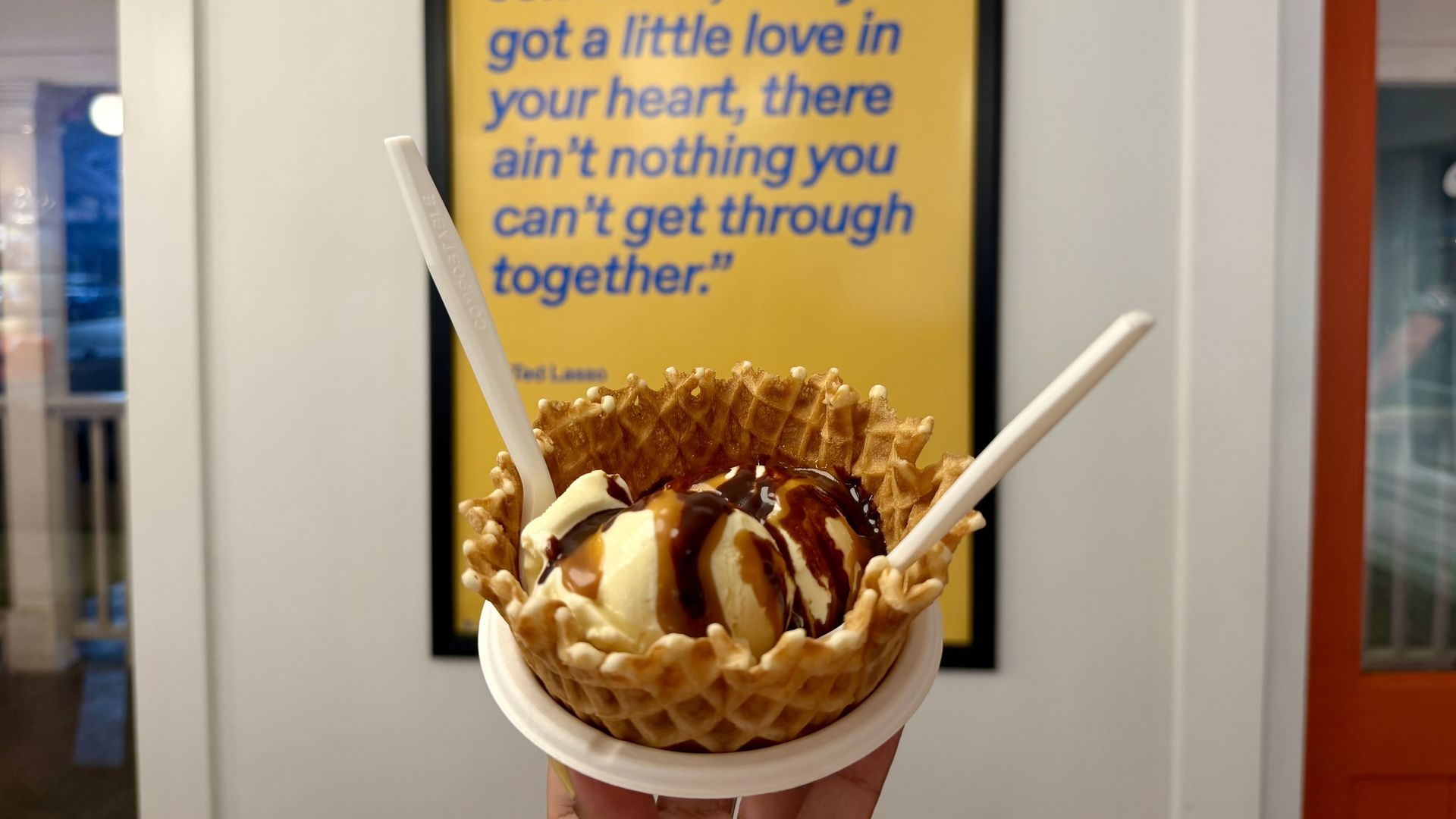 Hand holds a waffle cone with vanilla ice cream, drizzled with chocolate and caramel, in front of a yellow framed poster with blue text.