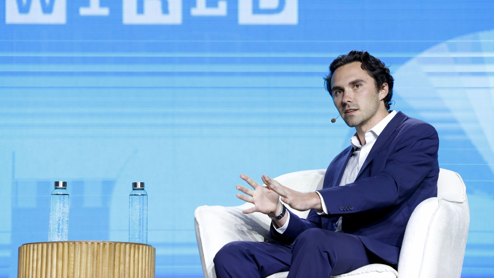 Man in a navy suit sits on a white armchair on stage, wearing a headset mic and gesturing with his hands. A wooden table with two water bottles sits to the left; a blue backdrop displays the word "WIRED".