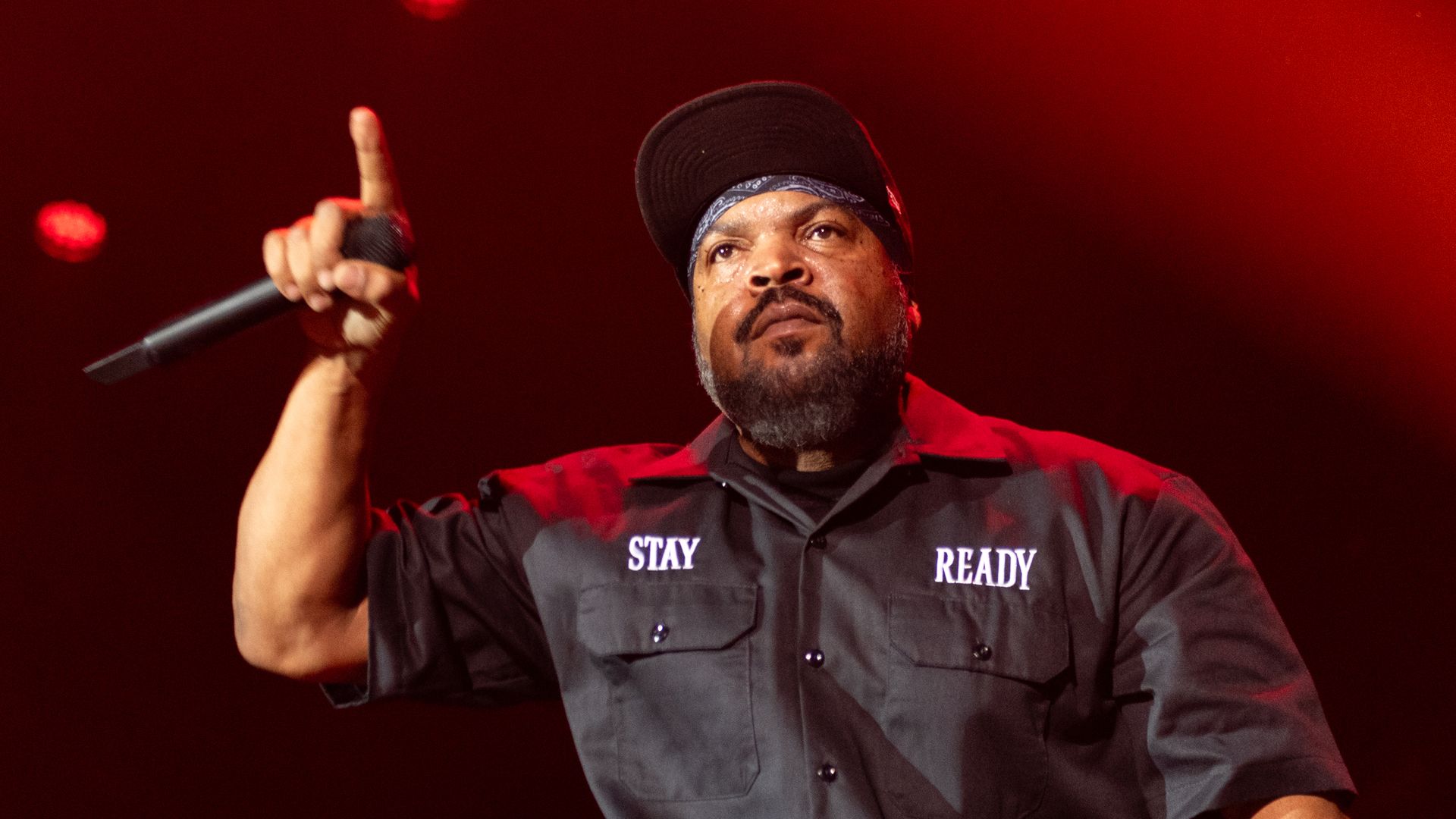 Ice Cube performs on stage against a red light and wearing a hat, with his hand holding a microphone in the air.