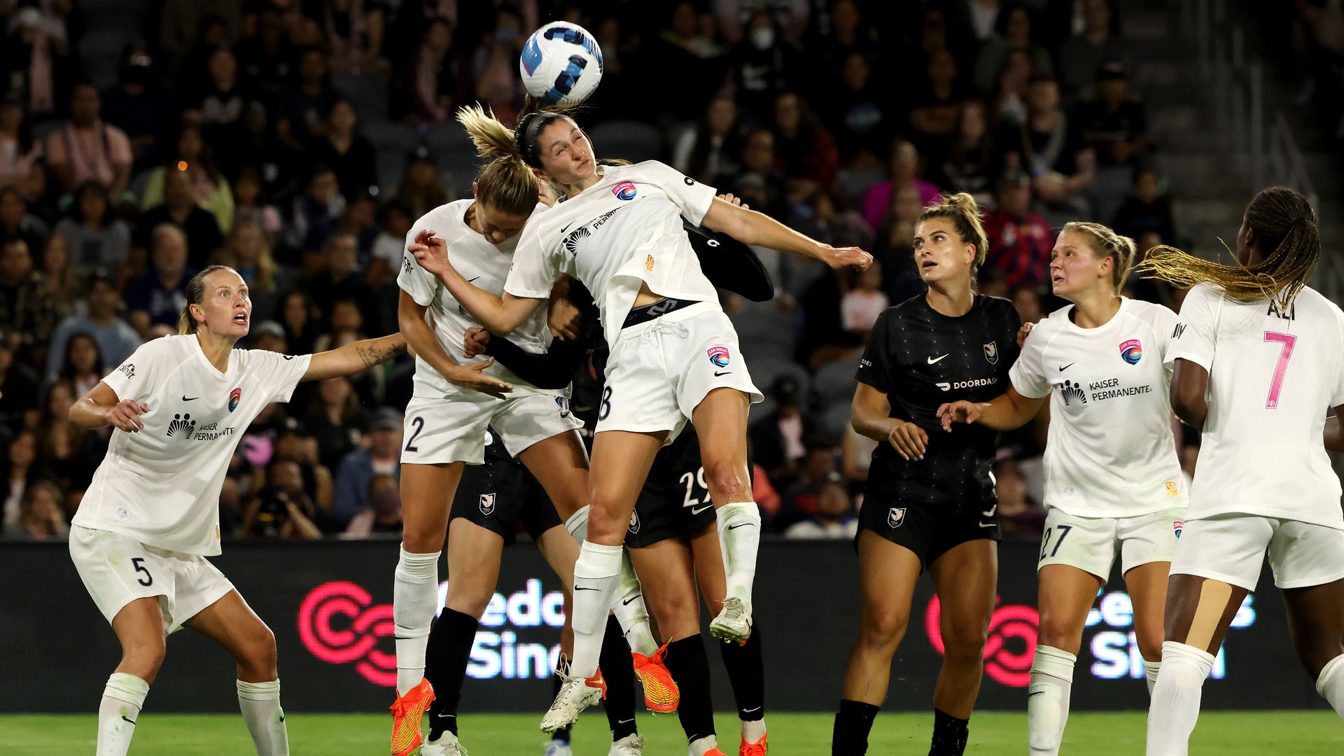 A woman in a white jersey wins a header against a crowd of women in black jerseys.