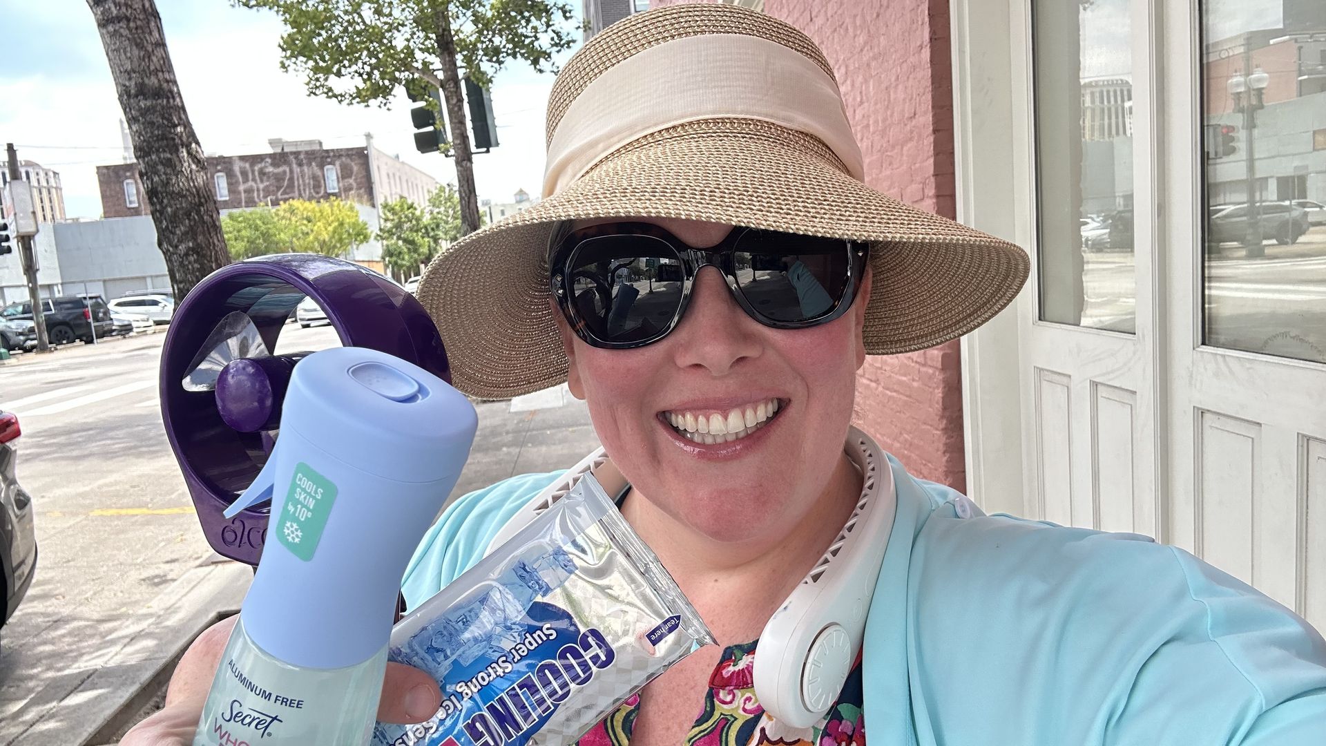 Selfie of a smiling woman in a wide straw hat and dark sunglasses on a sunny city street, holding a blue spray bottle and a neck fan.