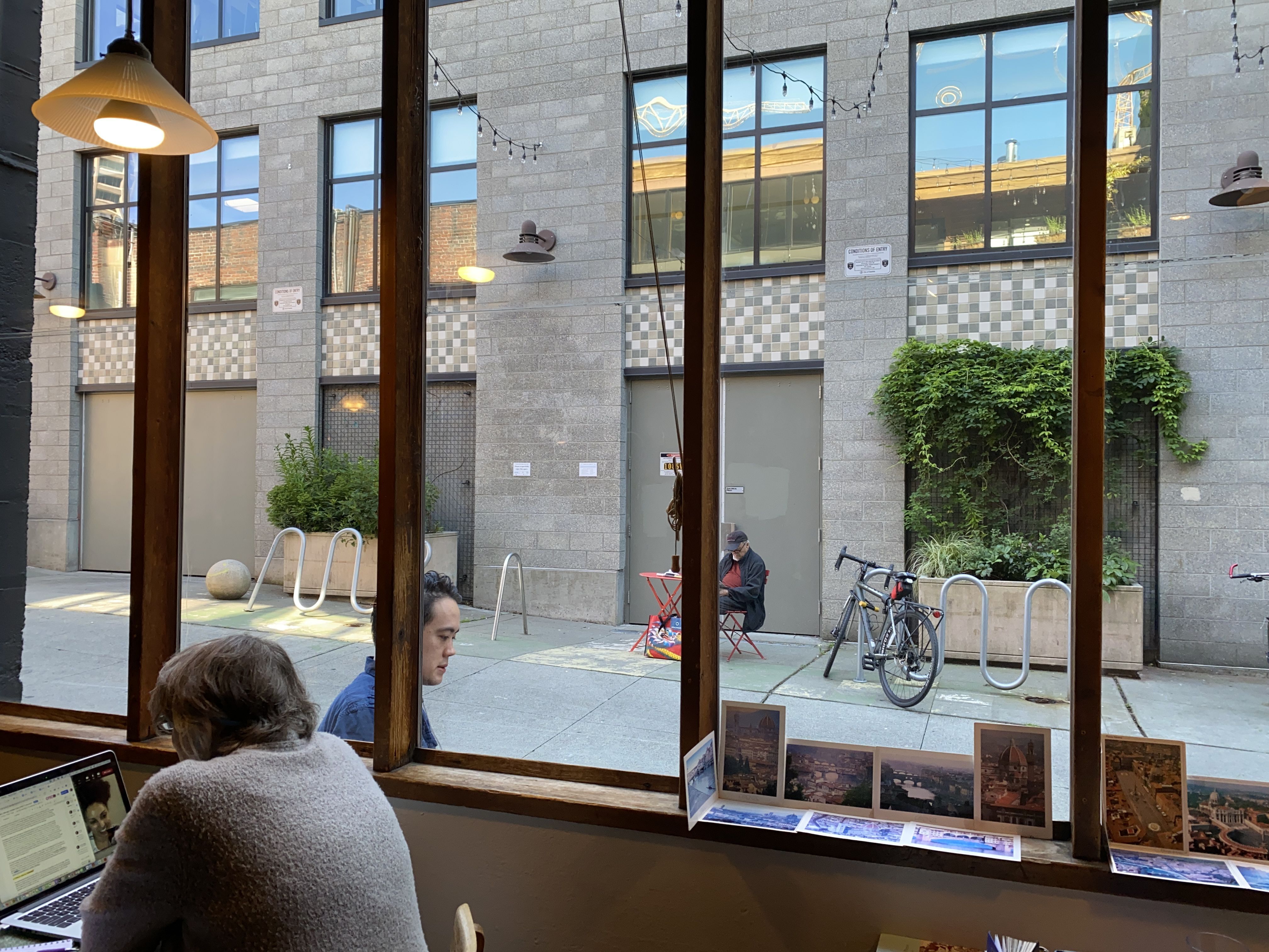 A man sits in front of large cafe windows with books on the windowsill and a view of a more modern building outside.