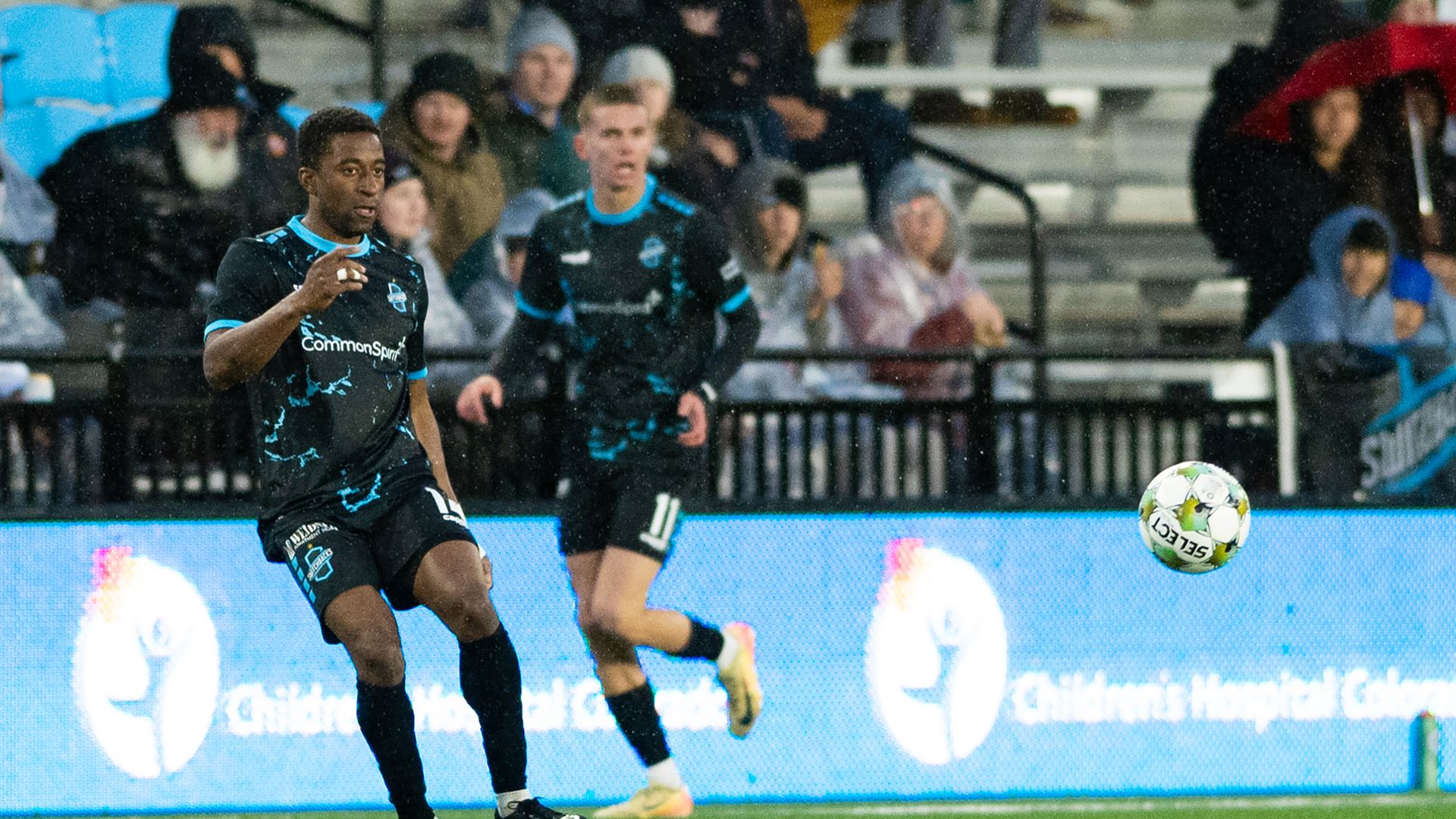 Two soccer players on a field watch a bouncing soccer ball. Both men are wearing the same uniform, black with light blue details. Behind them is a blue field barrier, and a few spectators sitting in stands. 