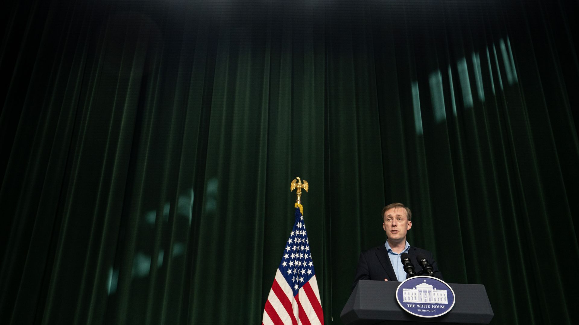 A man stands behind a White House podium with microphones, next to a U.S. flag with a gold eagle finial, against dark green curtains under bright stage lights.