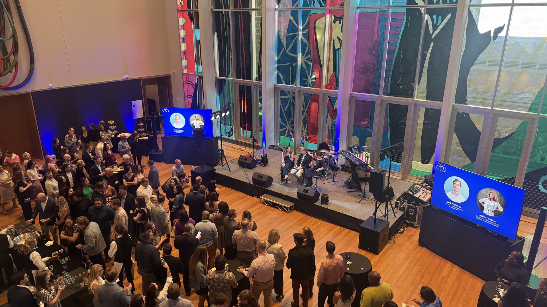 Large indoor event with diverse audience watching a panel discussion on stage with three seated speakers. Two large screens show speaker photos and names, colorful stained glass windows in background.