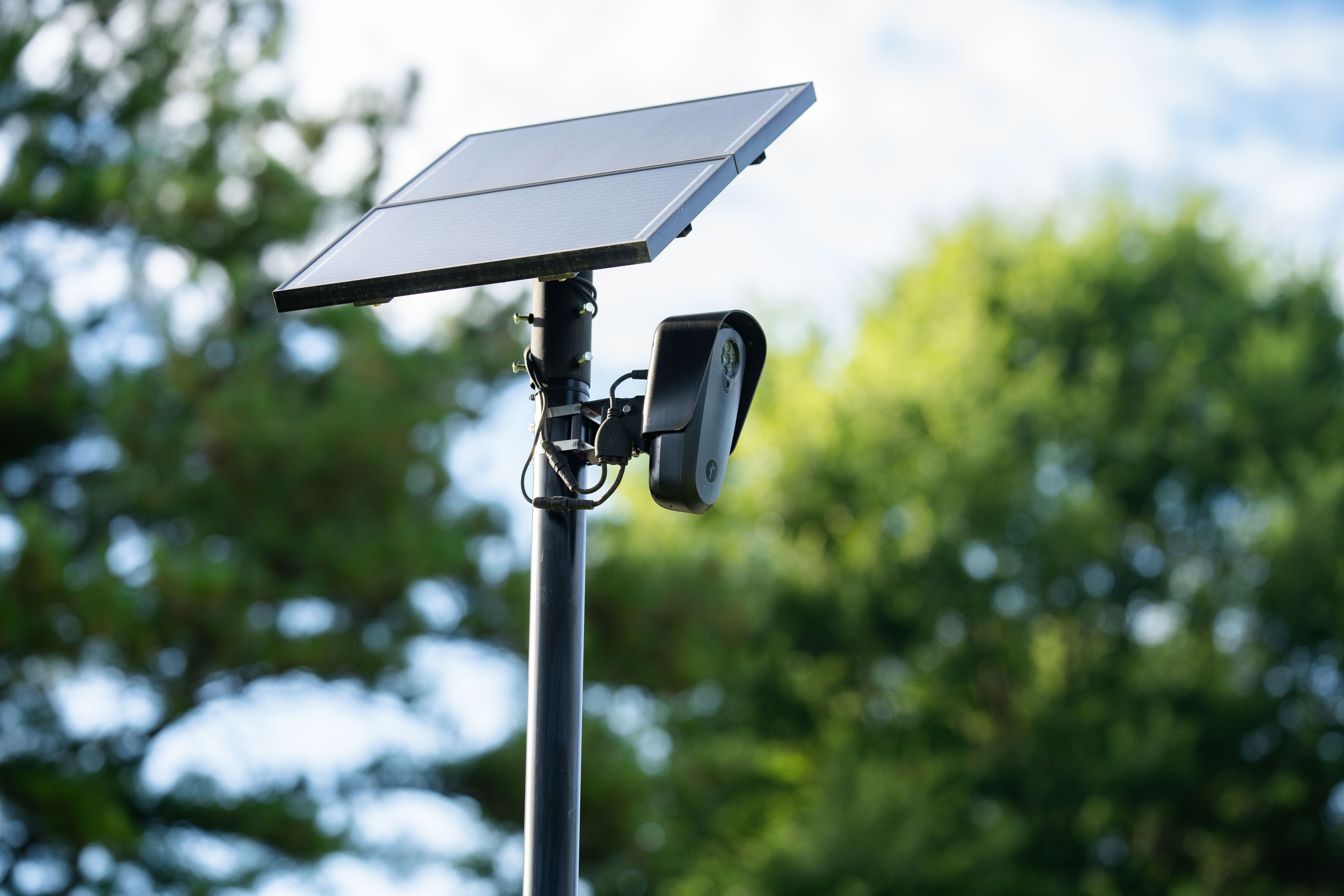 Solar panel array mounted on a pole with a license plate reading Flock camera housing beneath, outdoors with a blurred green treeline and blue sky in the background.