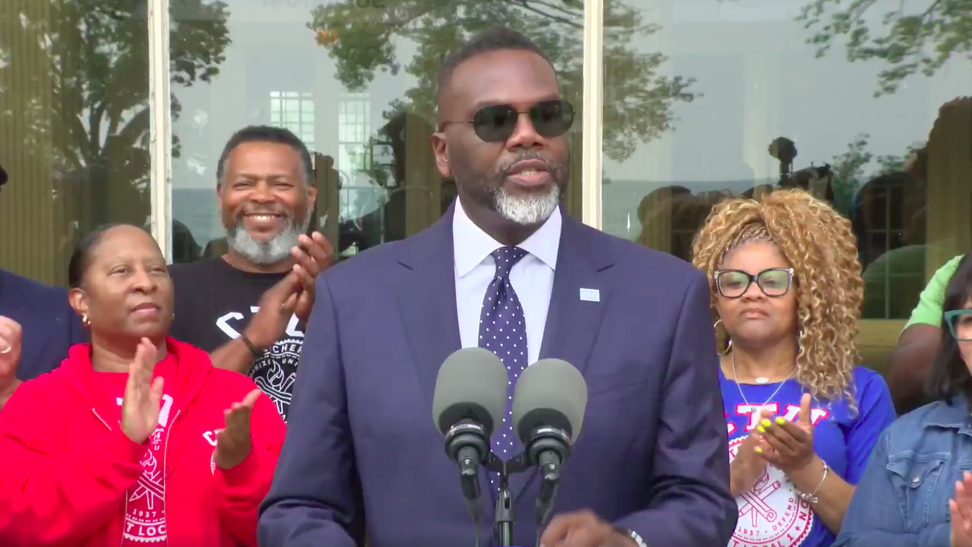 A man in a navy suit, white shirt, polka dot tie, and sunglasses speaks at a podium with two microphones. People behind him clap, wearing casual clothing and glasses.