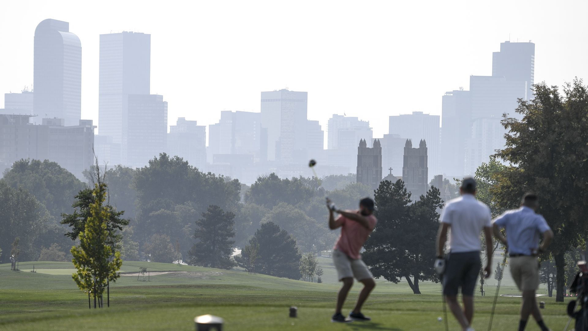 A man prepares to swing a golf club while two other man watch during a sunny but hazy day at a golf course. 