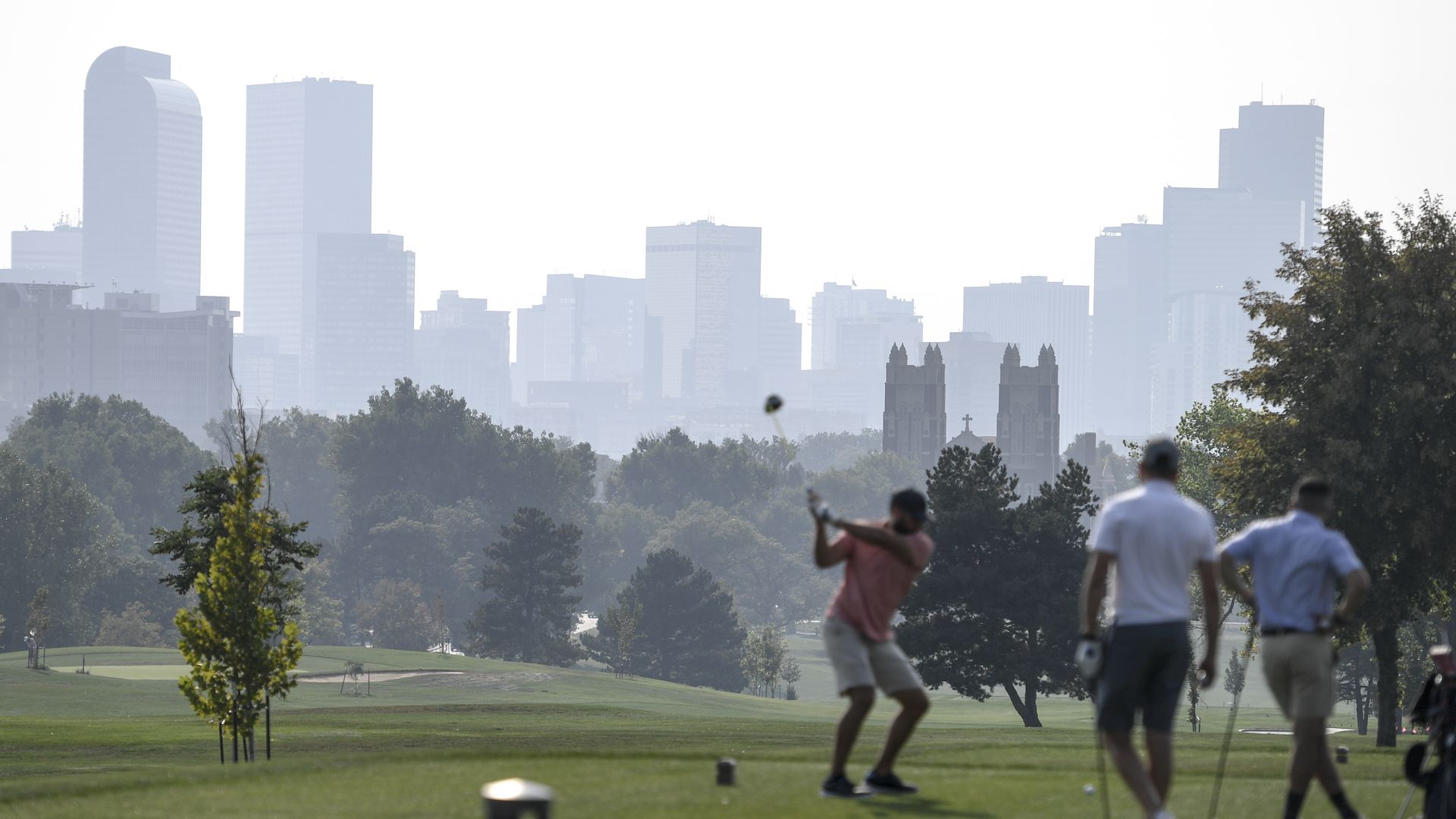 A man prepares to swing a golf club while two other man watch during a sunny but hazy day at a golf course.
