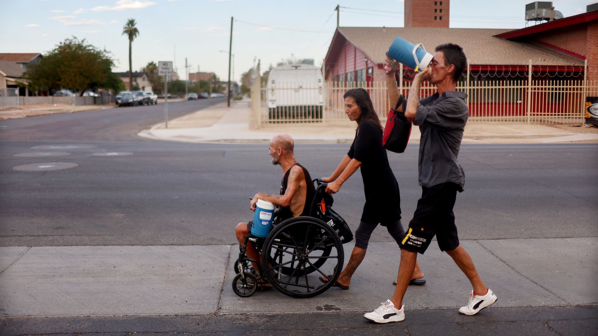 Three people cross a city street: a man in a wheelchair at the front, a woman walking beside him, and a man holding a blue water jug to his mouth; red-brick buildings and a fence behind them.