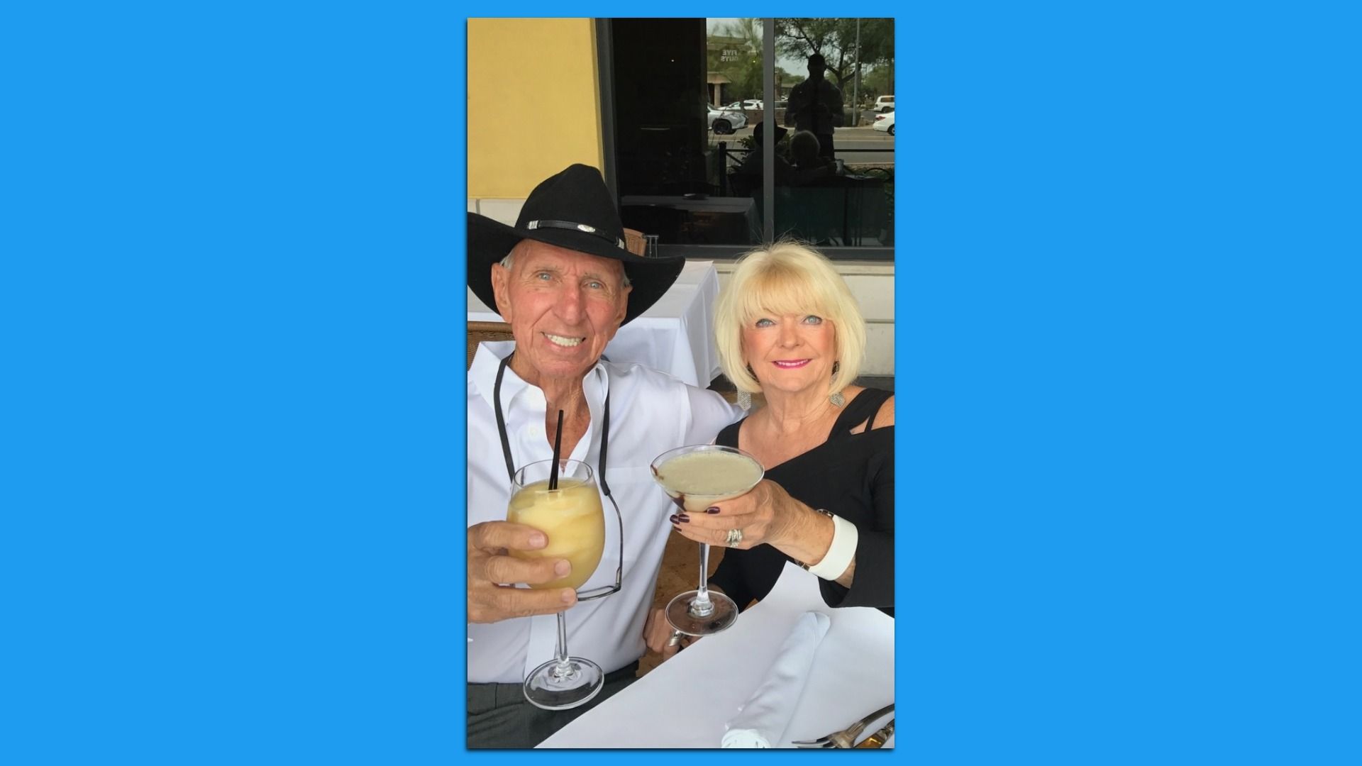 Smiling elderly man in black cowboy hat and white shirt and woman with blonde hair in black dress hold cocktails at a restaurant table with white tablecloth and napkins.