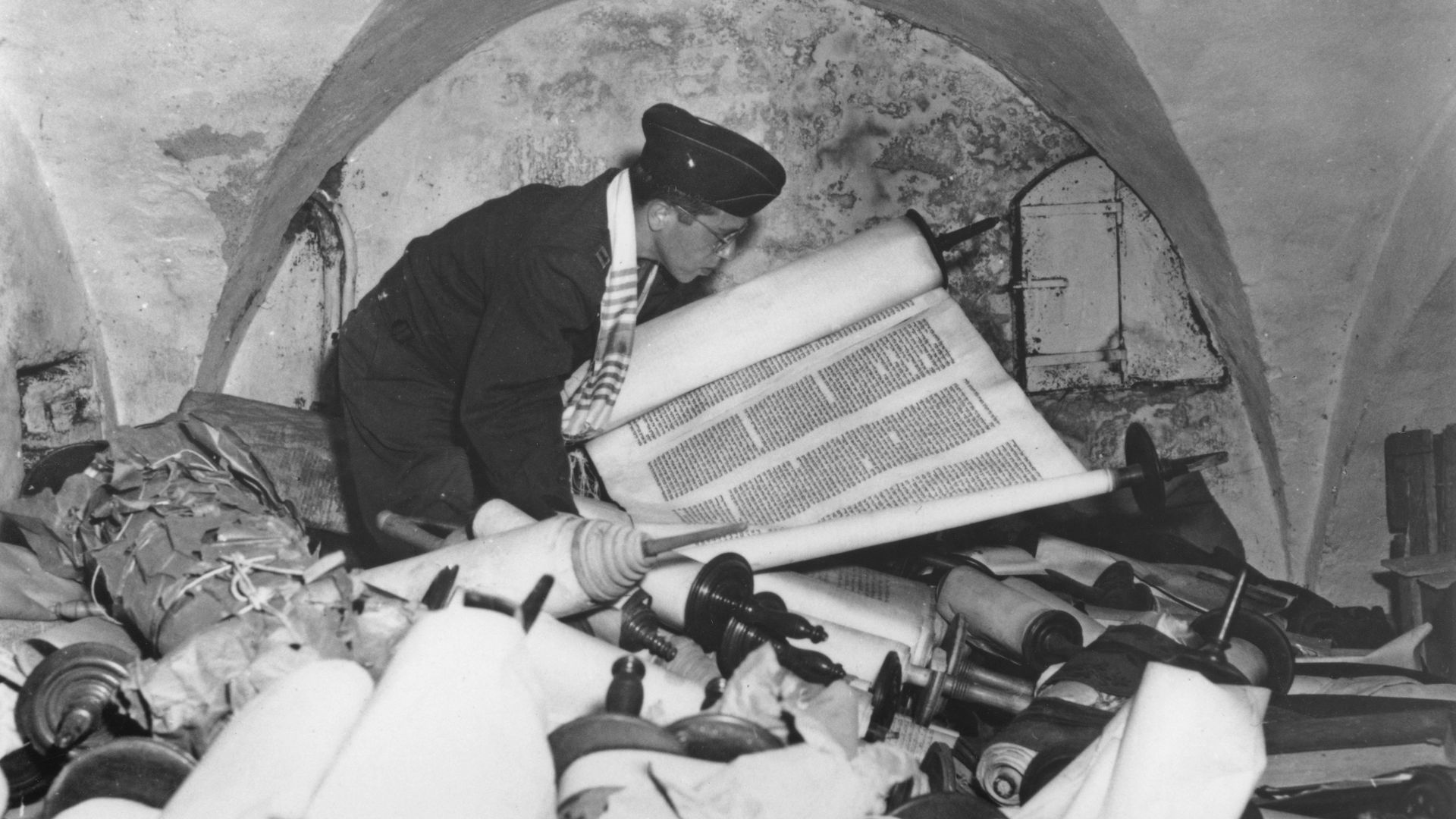 Black and white archival photo of a US army chaplain examining a torah scroll (amidst a pile of scrolls)