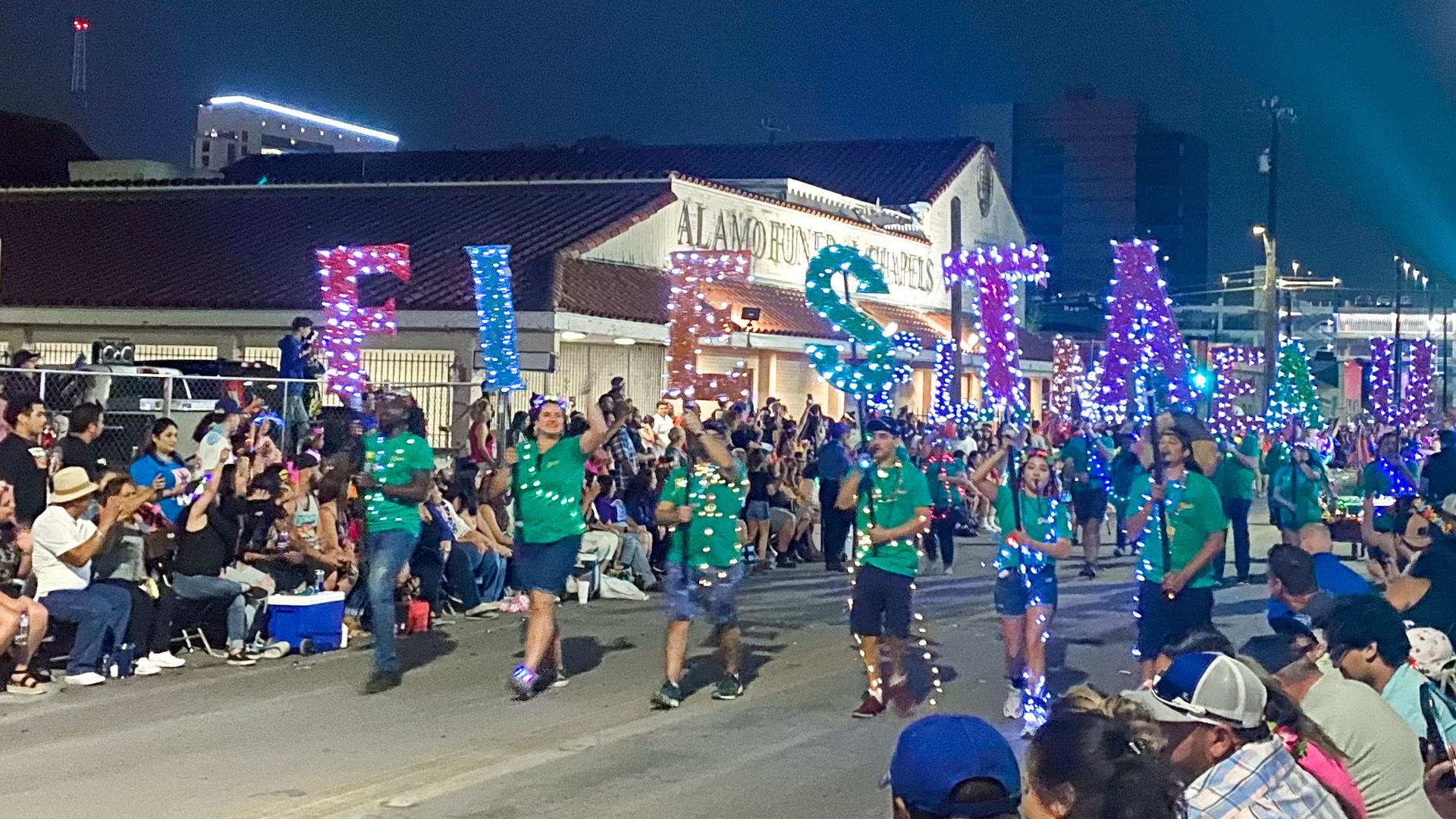 Parade participants walk the route holding multi-colored letters that spell "Fiesta."