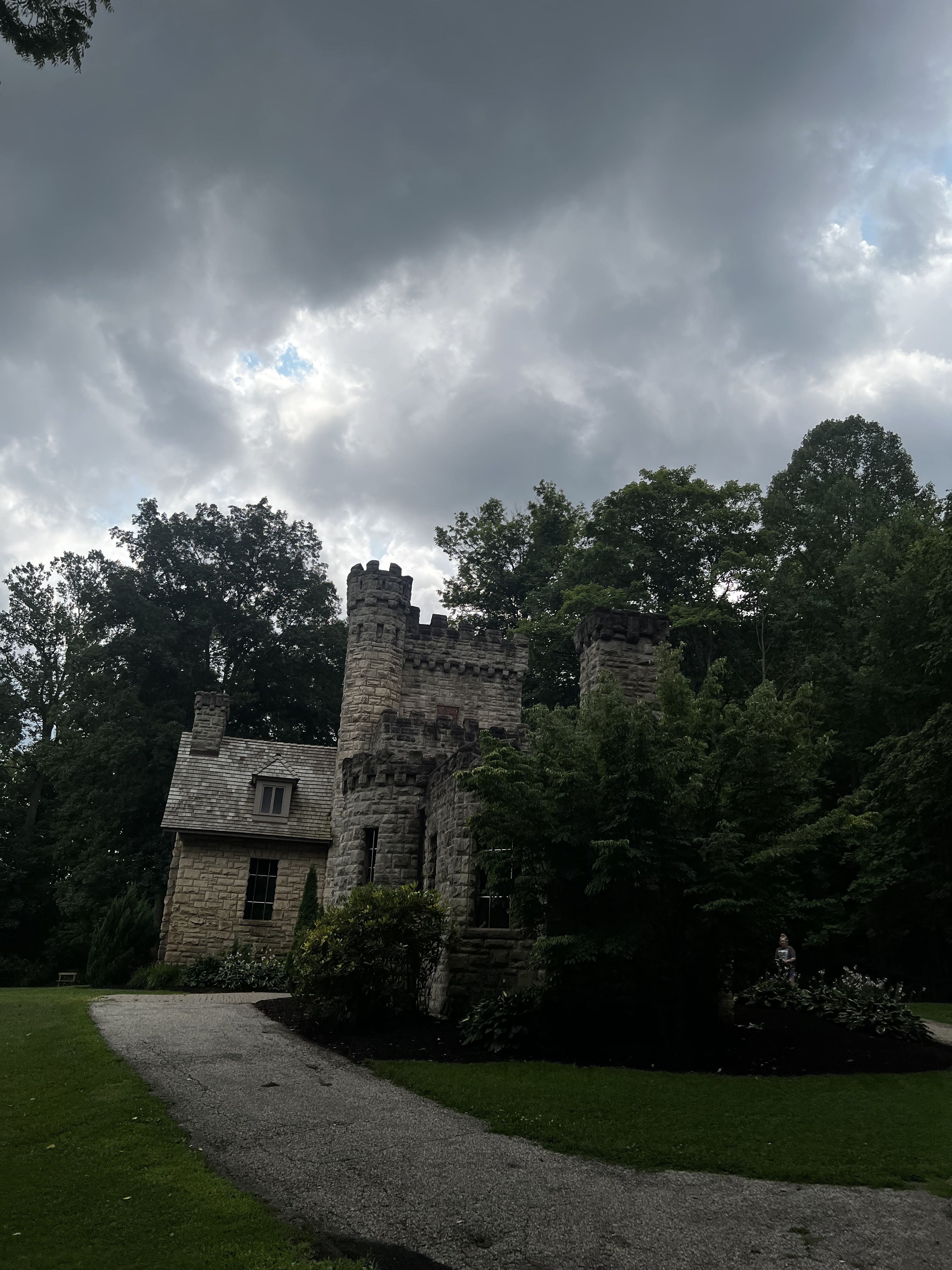 Stormy gray clouds hover above a stone castle ruin with crenellated towers, surrounded by dense trees. A gravel path curves through a manicured lawn in the foreground, with a small figure at the right.