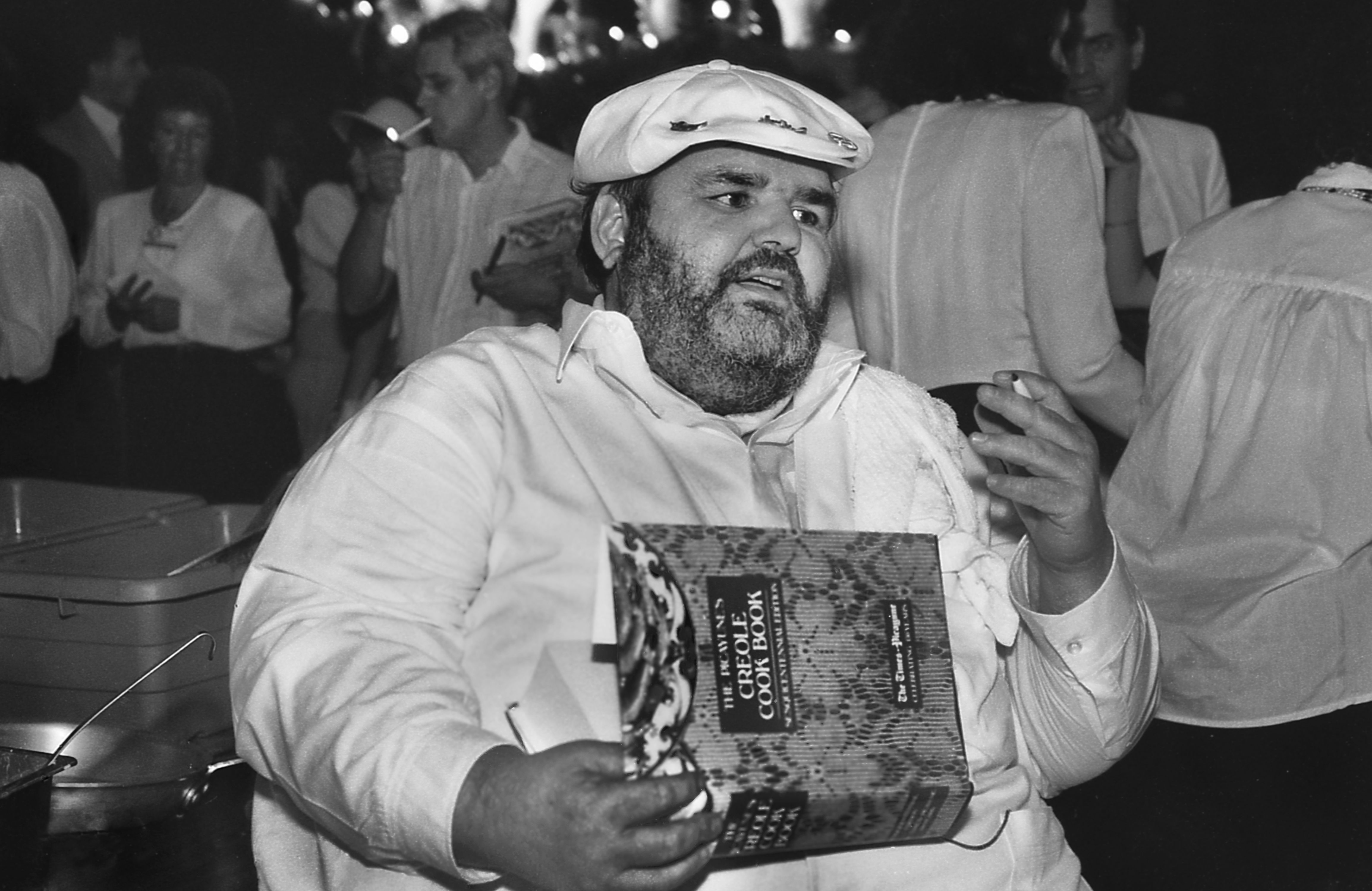 Photo shows chef Paul Prudhomme holding a cookbook at an event