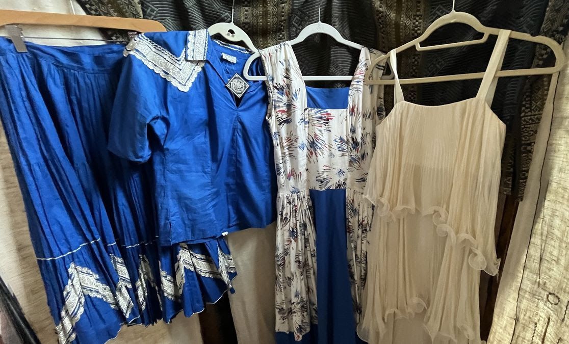 Four dresses hanging on hangers: a blue skirt and top with white lace, a blue and white floral dress, and a cream-colored dress with ruffled layers, set against a dark patterned background.