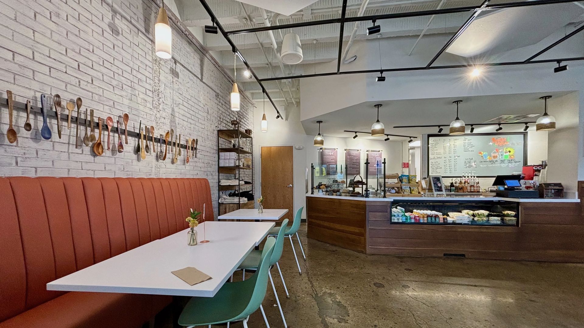 Modern cafe interior with white brick wall, wooden spoons hanging, rust-colored bench seating, white tables, mint green chairs, pendant lights, and a counter displaying food and drinks under bright lighting.