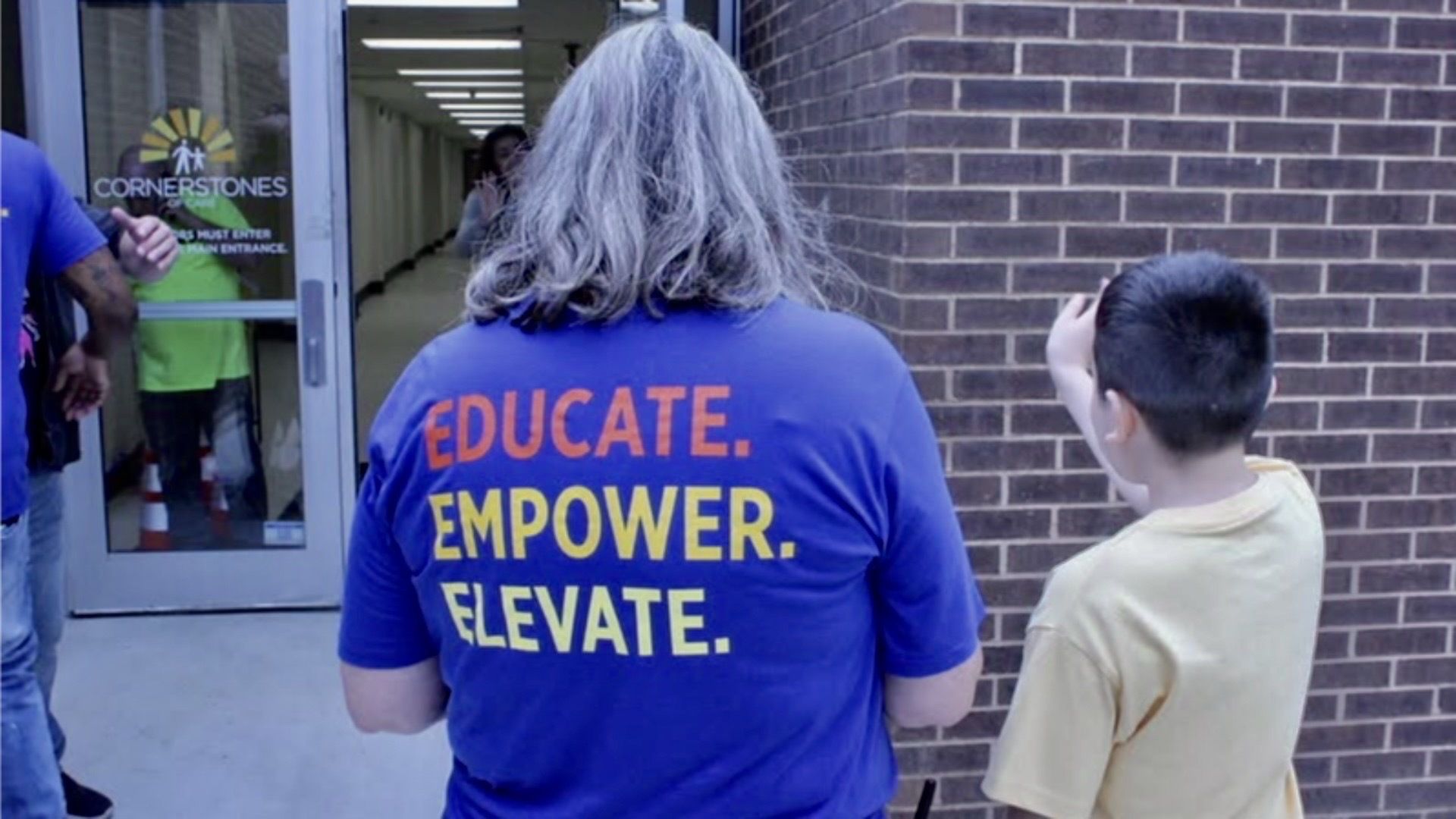 Back view of a person with gray hair wearing a blue shirt that says EDUCATE. EMPOWER. ELEVATE., standing beside a child outside a brick building with a glass door labeled CORNERSTONES.