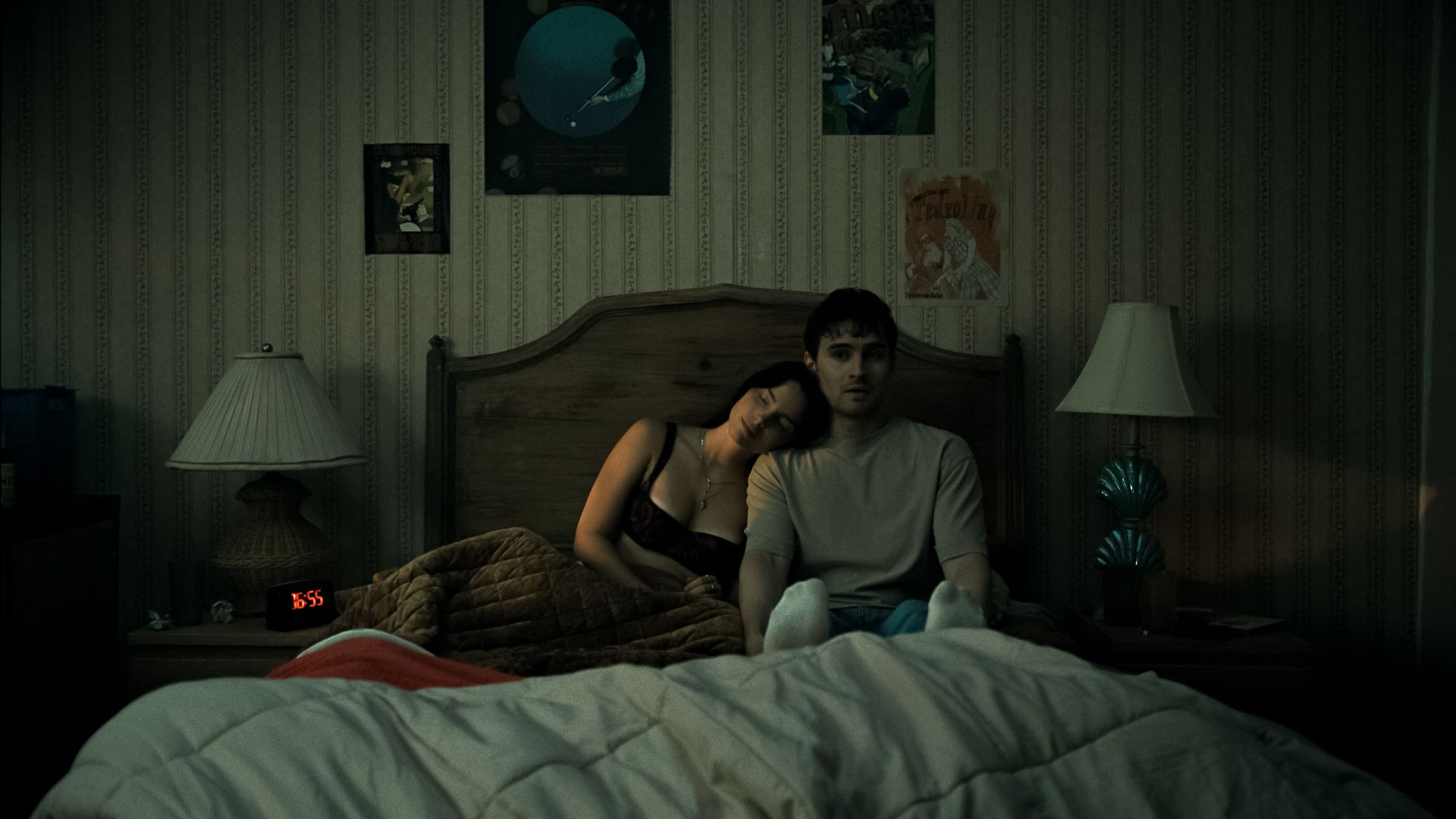Dimly lit bedroom scene with a couple sitting on a bed; the woman leans on the man. Vintage striped wallpaper with posters, two bedside lamps, a turquoise lamp on the right, and a red digital clock reading 16:55.