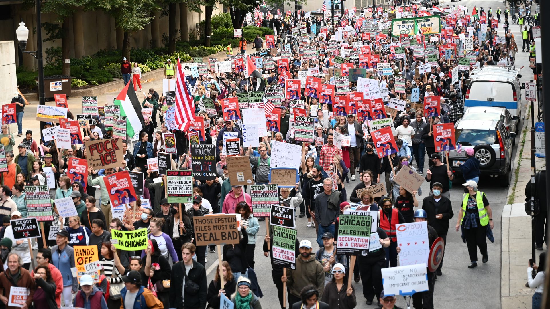 A dense city street protest with hundreds marching, holding bright signs and flags (Palestinian and American). Buildings and parked cars line the avenue.