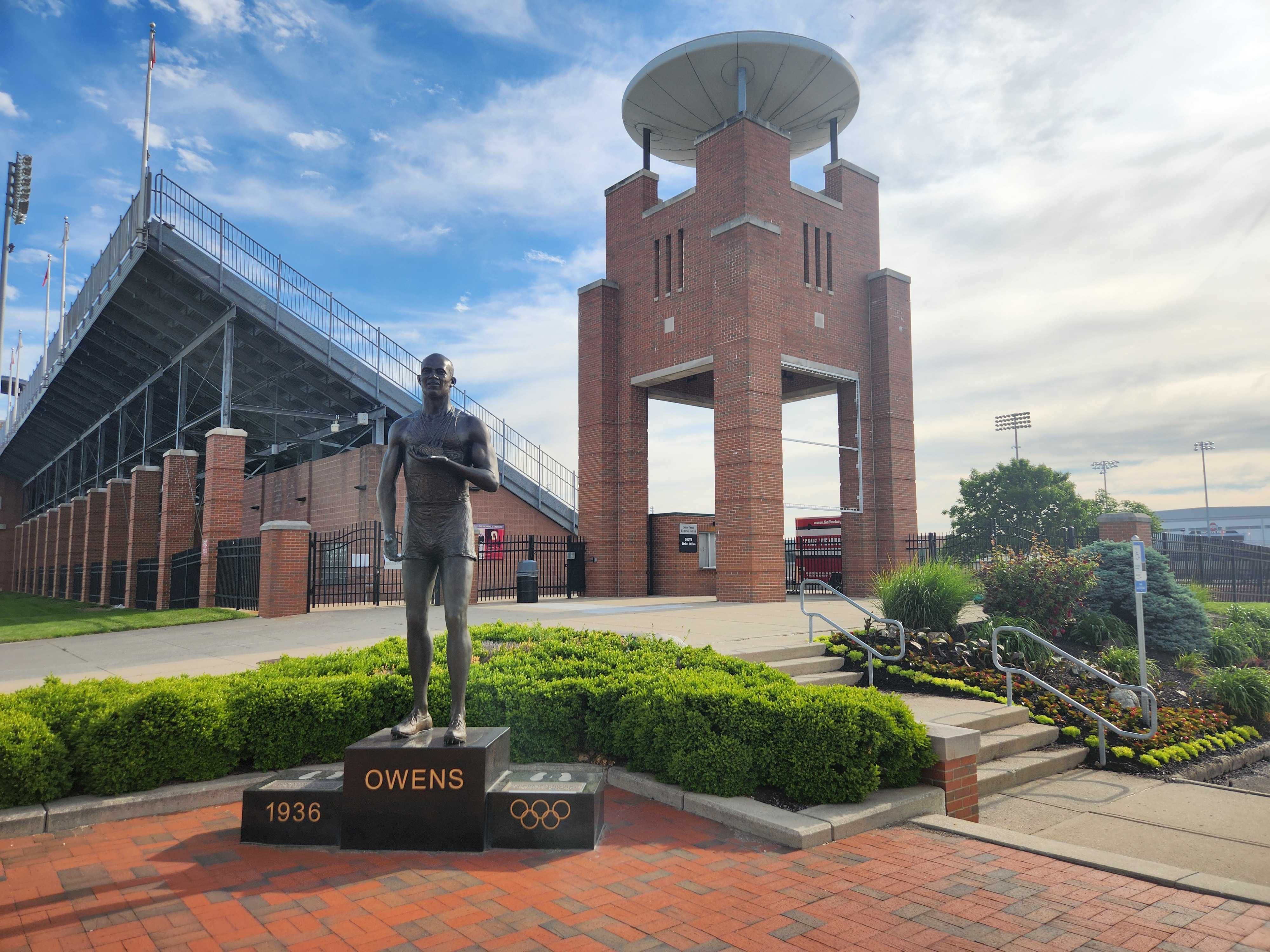 Bronze statue of Jesse Owens stands in front of a brick stadium tower with a circular rooftop. The pedestal shows OWENS, 1936, and Olympic rings; bleachers and flags sit under a blue sky.