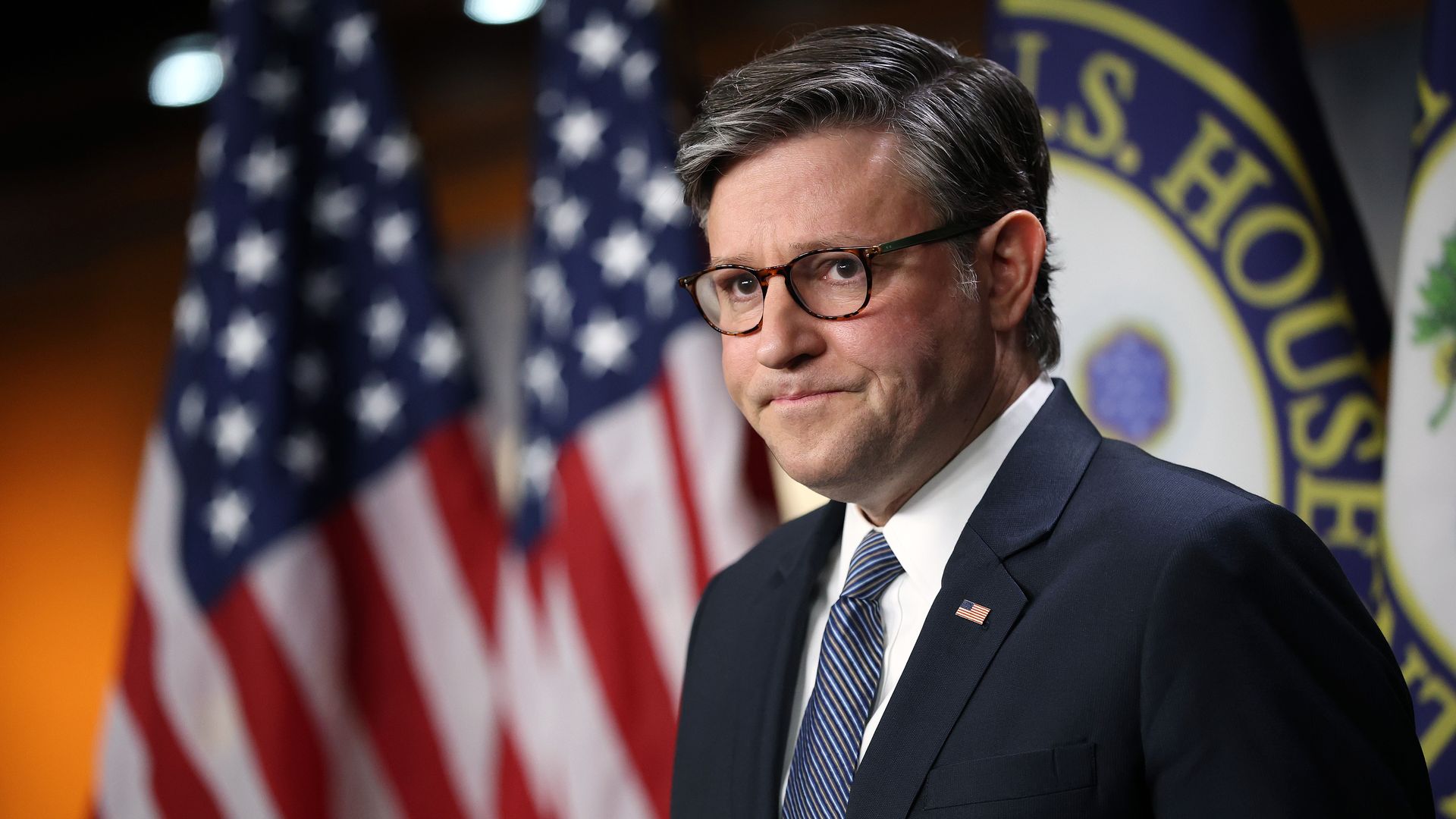 A man in a dark suit, striped blue tie, and tortoiseshell glasses stands before American flags and a blue-yellow banner, wearing a small US flag pin and smiling.