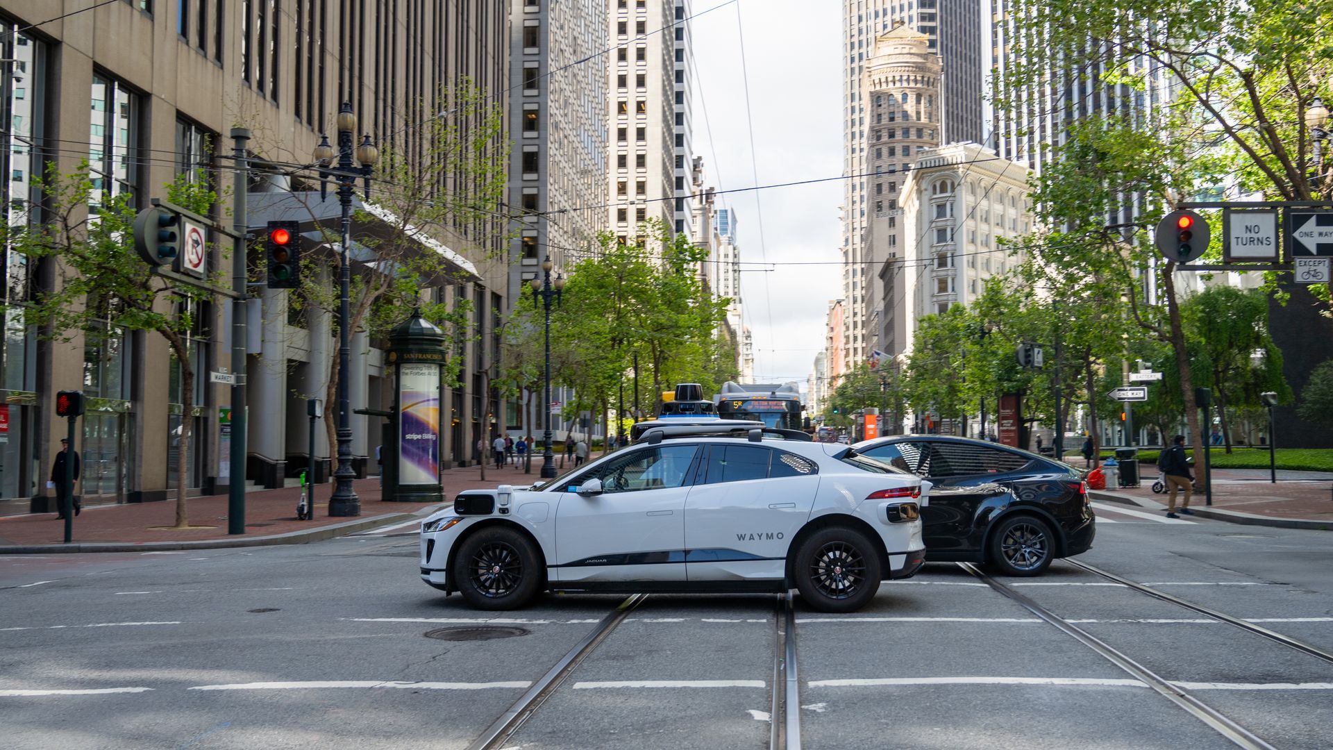 Urban street scene with tall buildings lining both sides, tram tracks in the center, overhead wires and traffic signals; foreground features a white Waymo autonomous taxi with roof sensors.