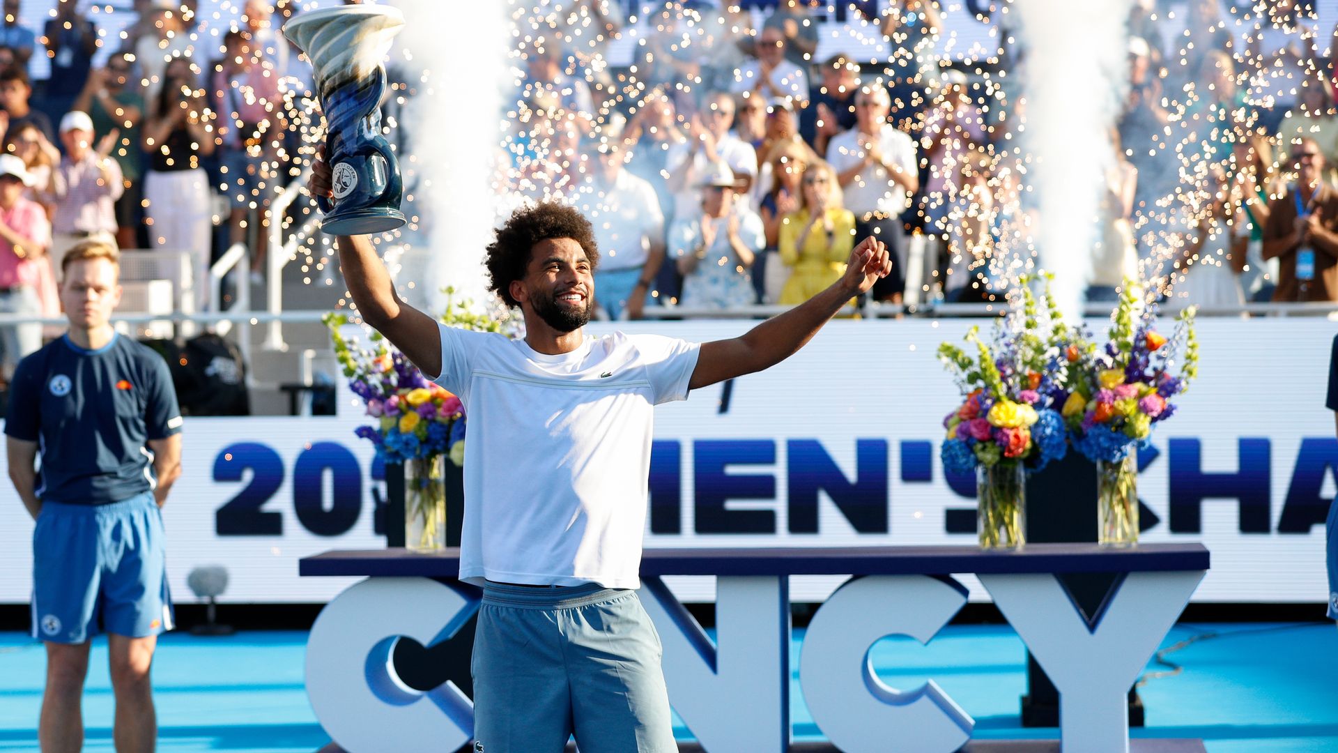A tennis player in a white T-shirt and short poses with a trophy in his right hand (left side of the picture) as a crowd in the background cheers for him. Confetti and smoke shoot out from a "Cincy: Cincinnati Open" pedestal behind him.