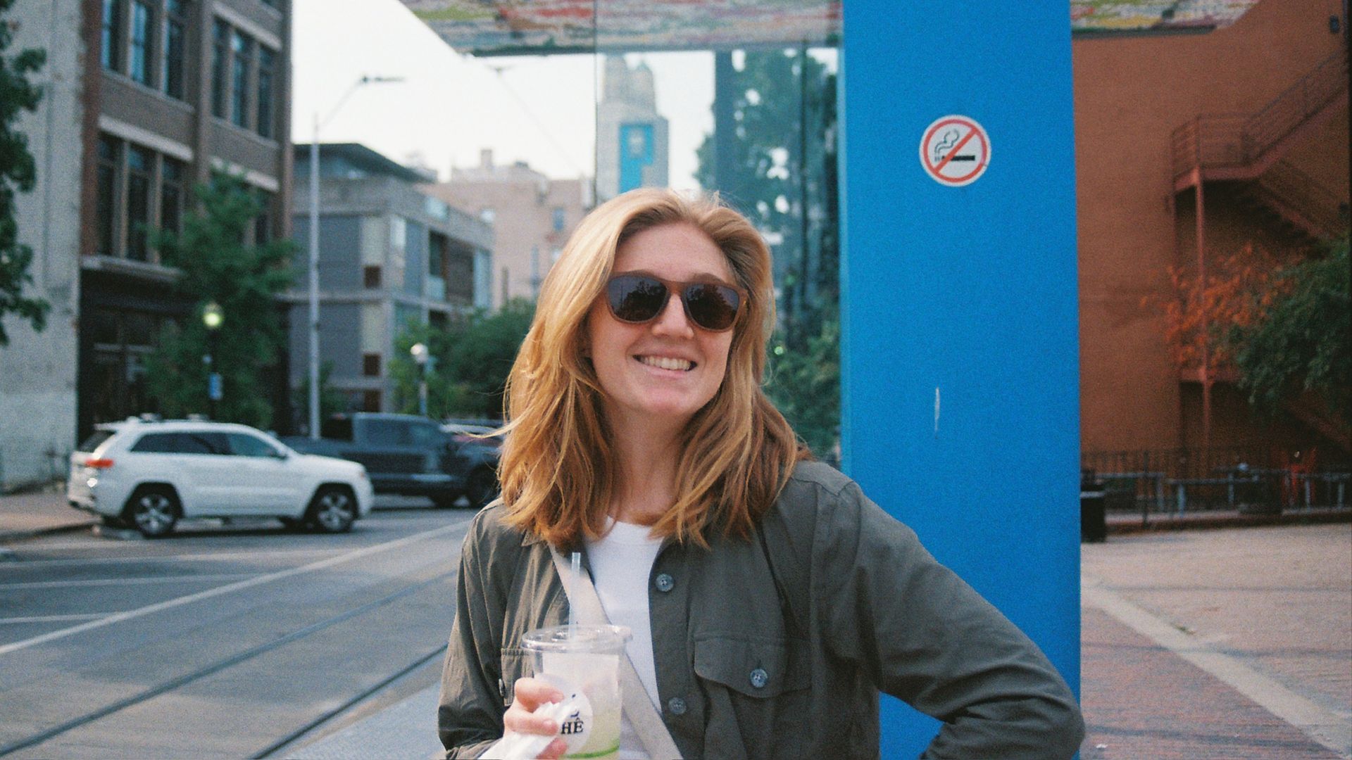 Smiling woman in sunglasses holds a drink beside a bright blue pillar on a city street; colorful overhead artwork, modern buildings, and a no-smoking sign are visible.