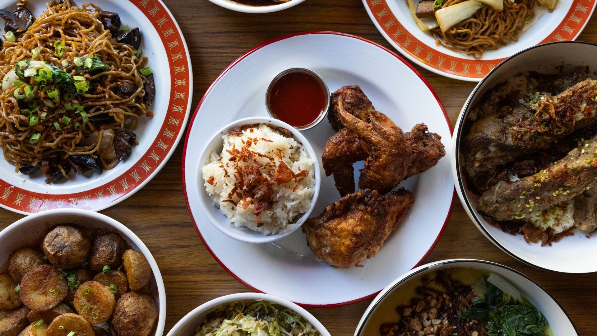 Variety of Asian dishes on a wooden table, including fried chicken wings with white rice, noodle dishes with greens, a stuffed roll in dark sauce, roasted potatoes, and vegetable salads.