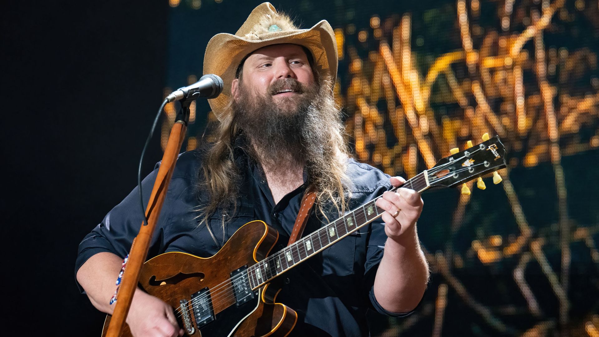 Chris Stapleton wears a straw cowboy hat while he stands at a microphone with a guitar. 