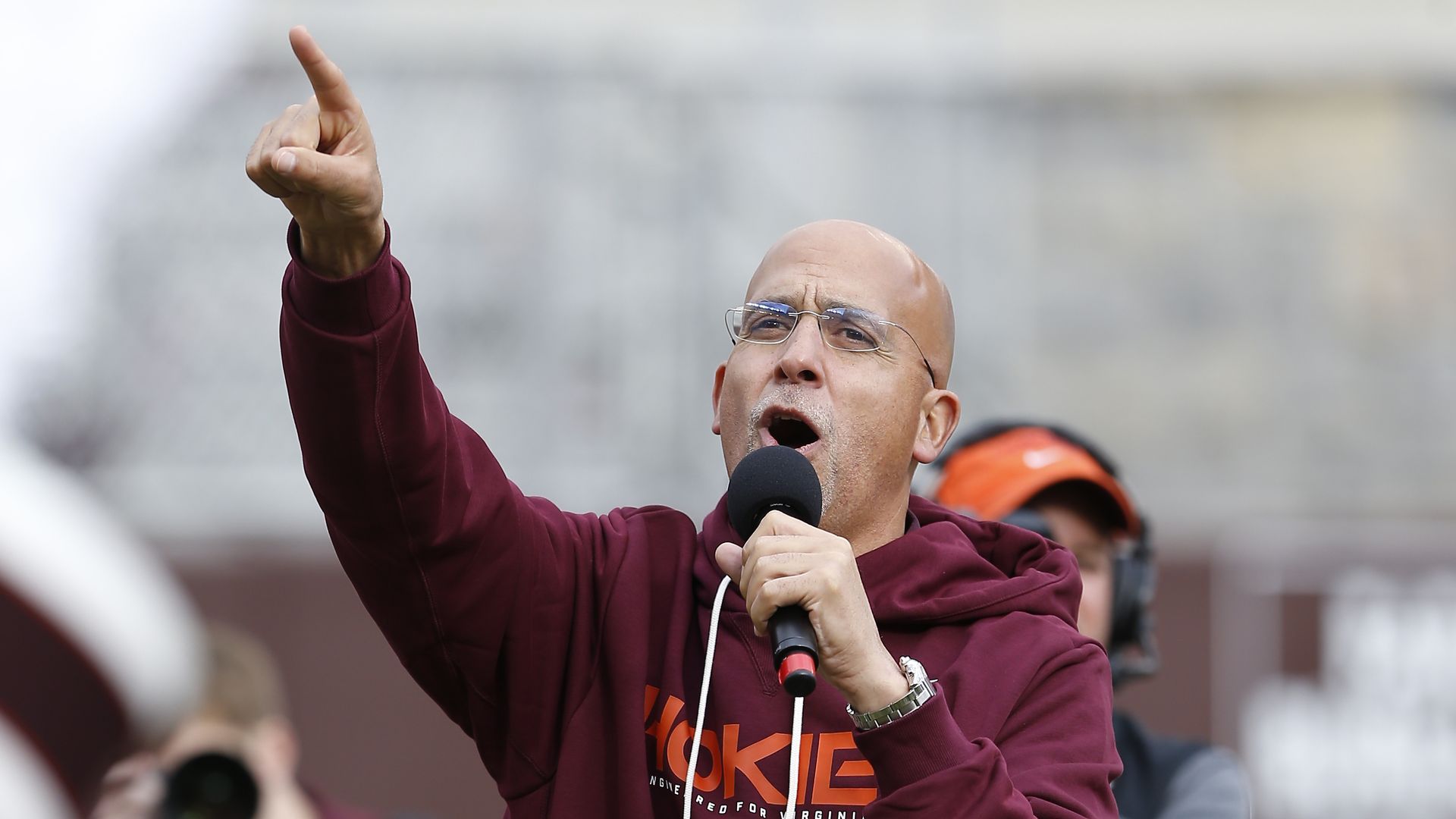 James Franklin with a mic in his hand and his right hand's index finger pointed up. He's wearing a Hokies sweatshirt while standing on a football field.