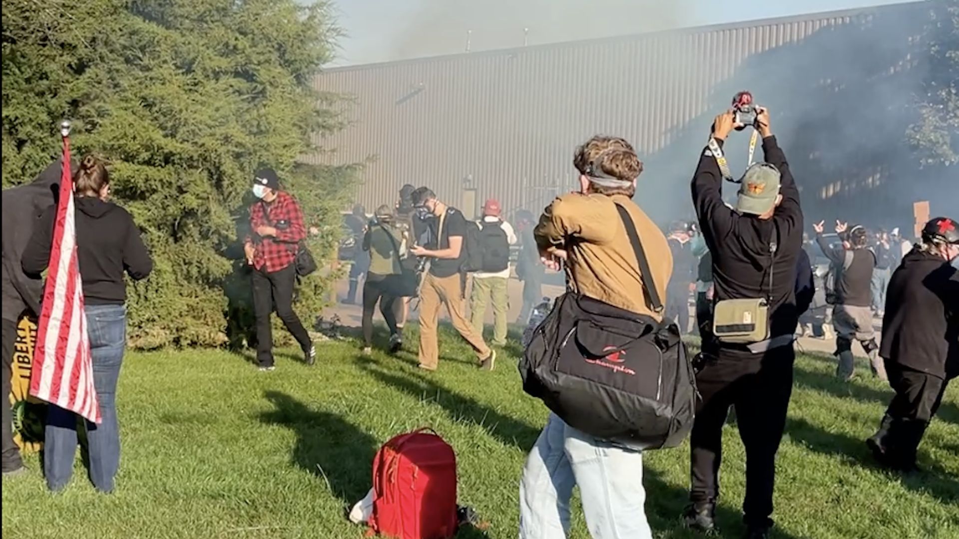 A group of people outdoors near a building, some wearing masks and carrying bags. One person holds a camera overhead, another holds a flag. Smoke or haze is visible in the background.