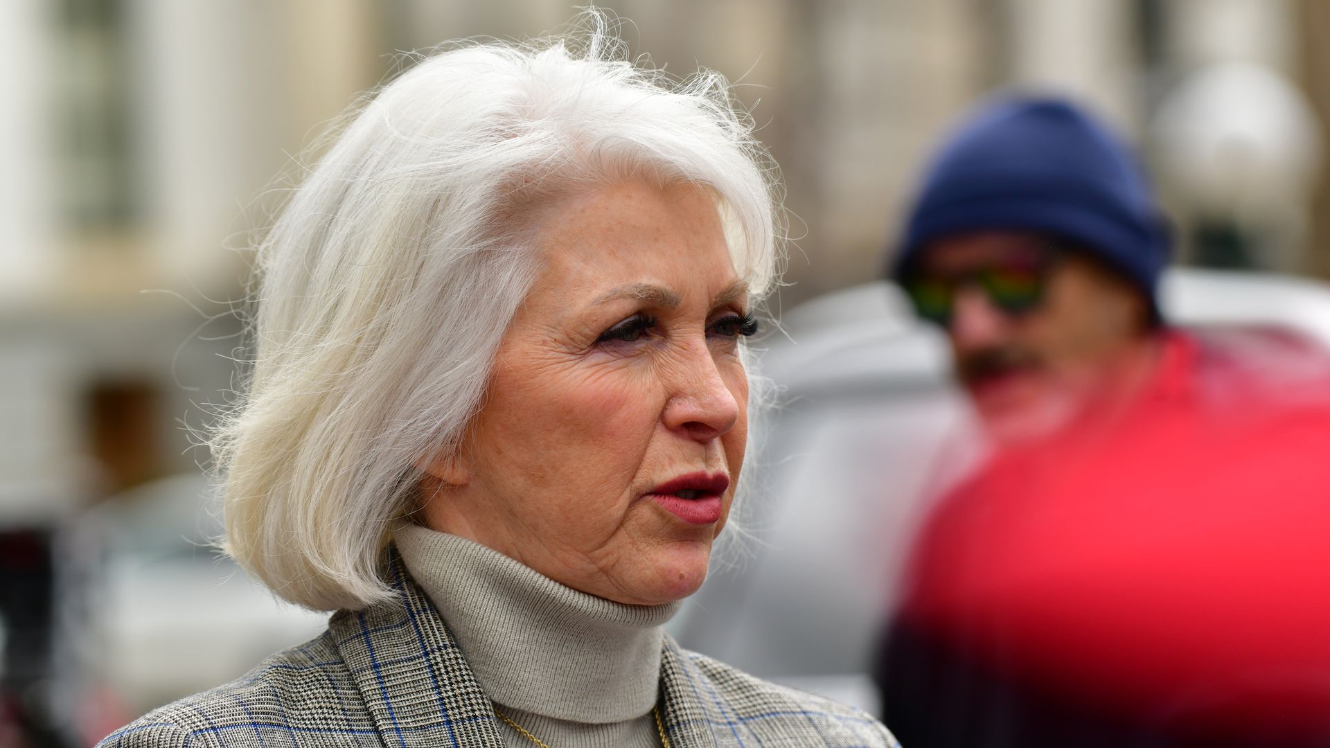 An older woman with short white hair wearing a light gray turtleneck and plaid blazer, looking to the right; a blurred man in a blue beanie and red jacket stands behind her outdoors.