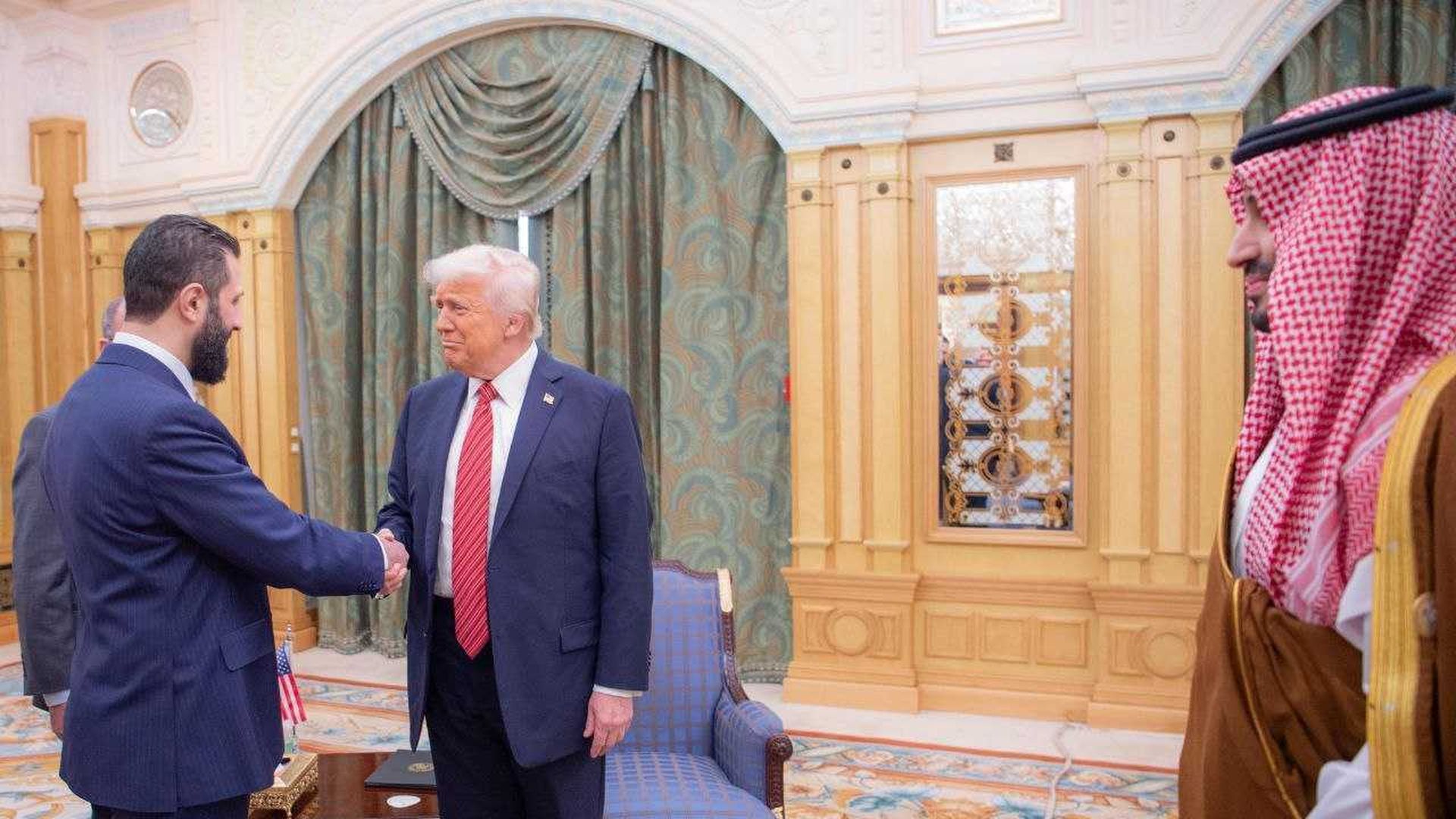 Man with white hair and red tie shaking hands with bearded man in blue suit in an ornate room with gold and green curtains; another man in red and white checkered headscarf watches.