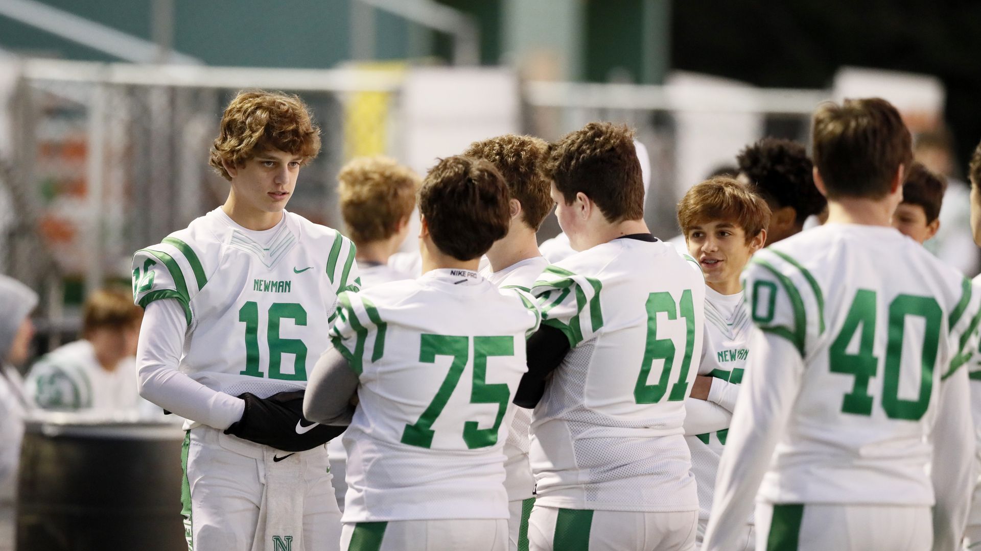 High school football players from Newman in white uniforms with green numbers, standing and talking on the sidelines during a game or practice at night.