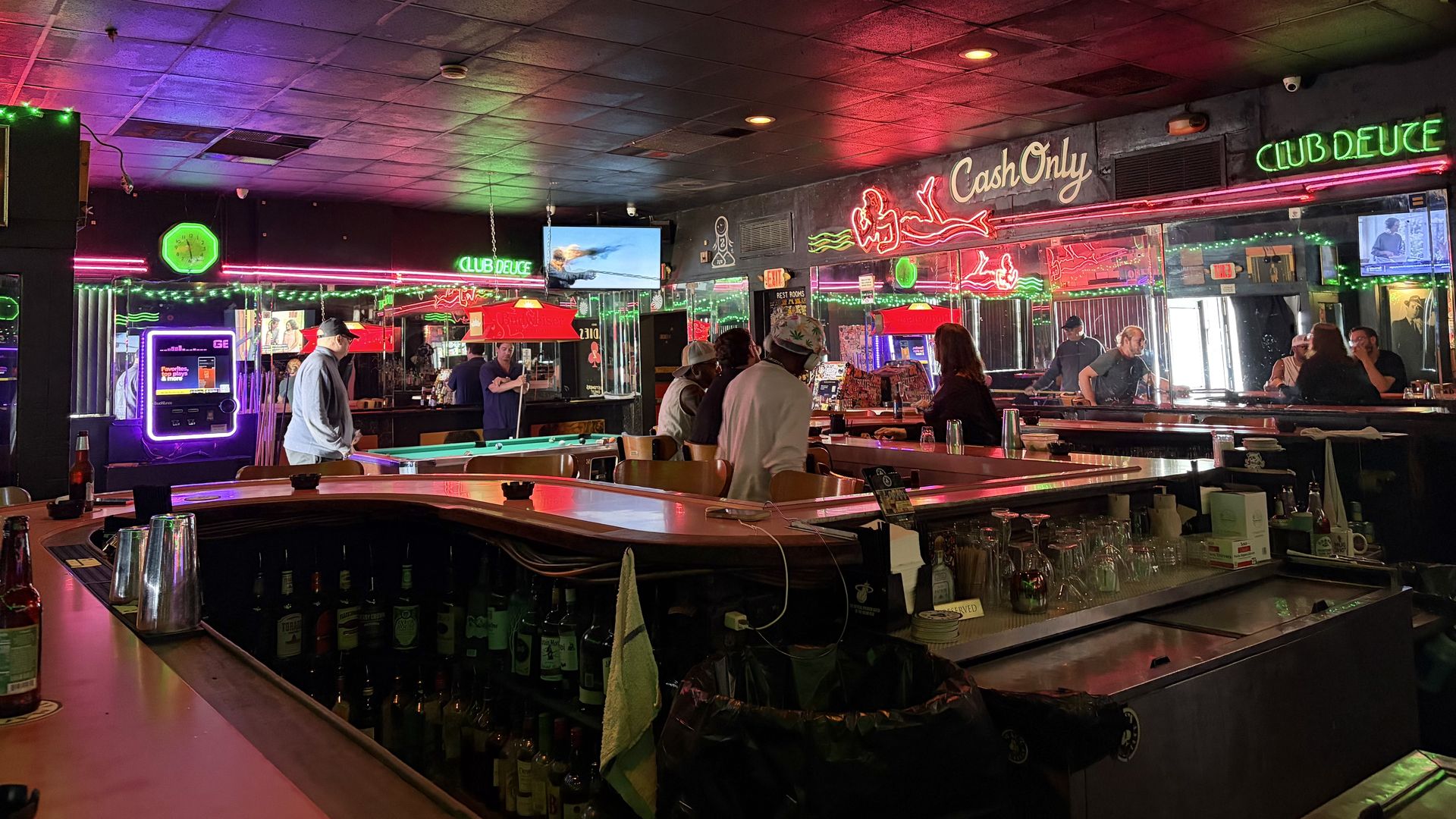 Neon-lit bar with pool tables, a long counter, and patrons. Green "Club Deuce" sign, red neon art, and a "Cash Only" sign on the back wall under purple-pink ceiling lights.