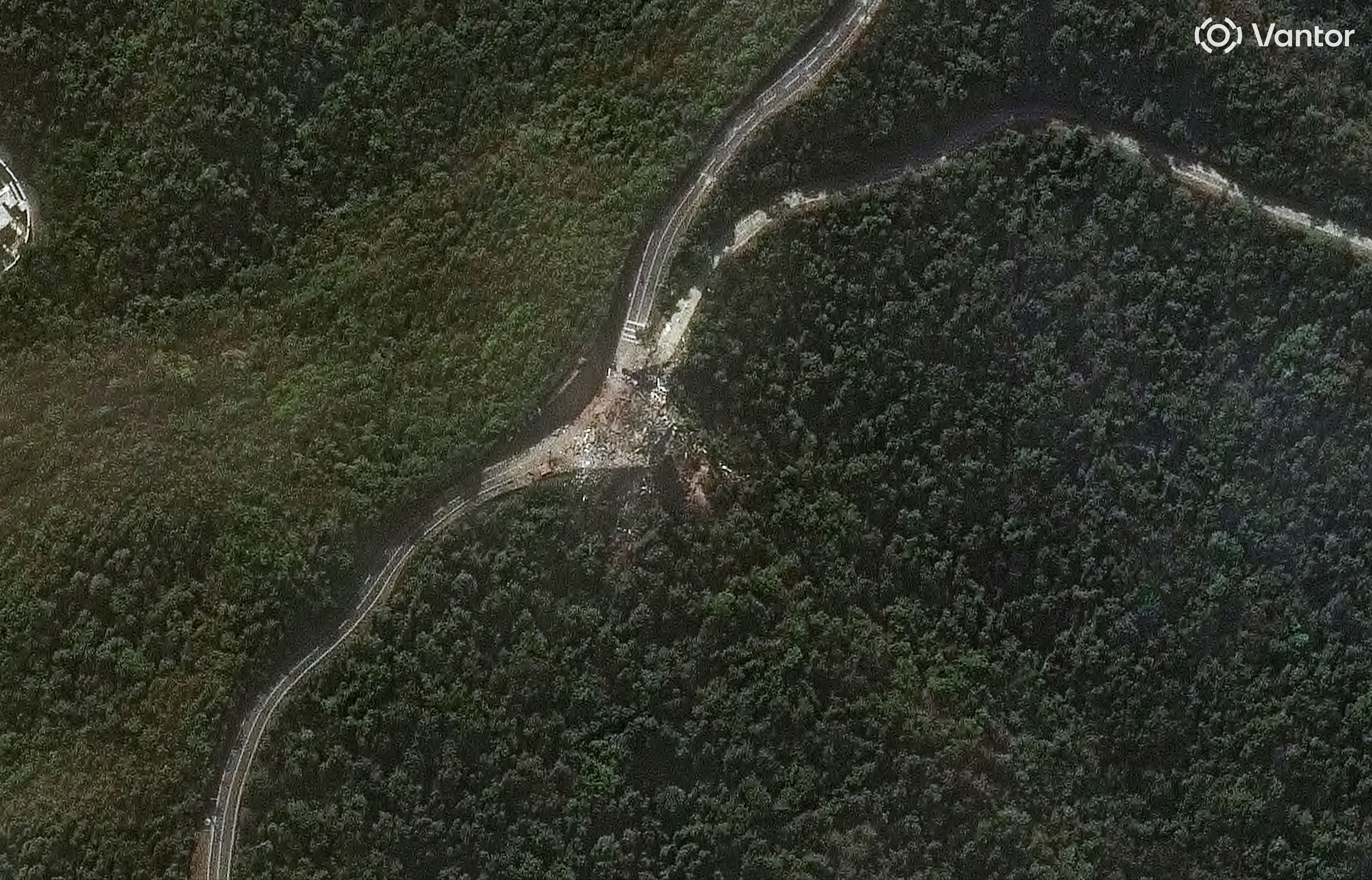 Aerial view of a forested area with a winding road intersecting. There is debris scattered at the road junction, surrounded by dense green trees.