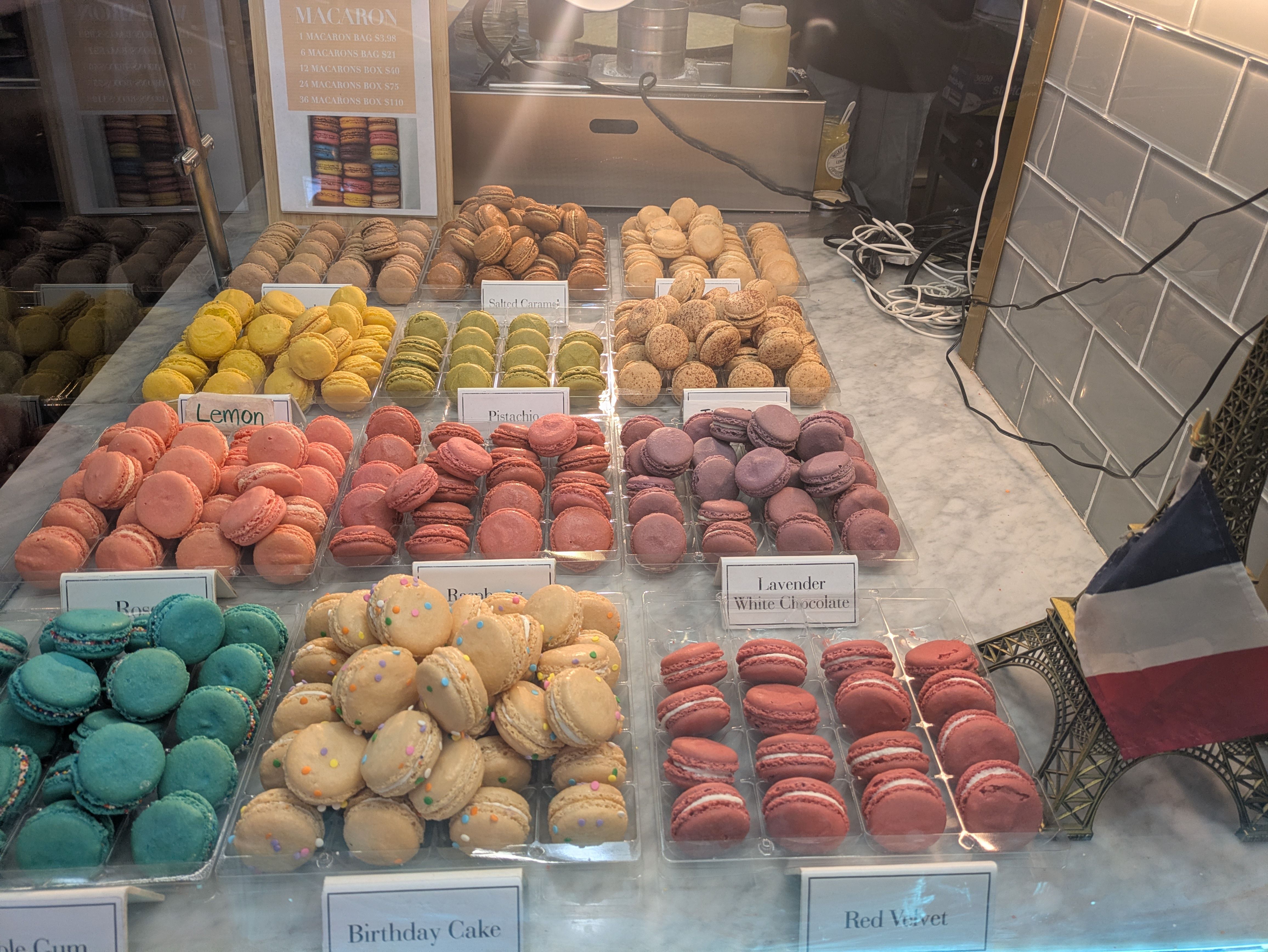 Display of colorful macarons in flavors like Lemon, Pistachio, Lavender White Chocolate, Birthday Cake, and Red Velvet on a marble counter with a small Eiffel Tower and French flag.