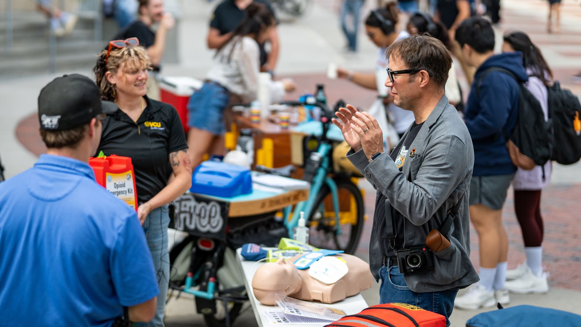 a crwod of people blurred in the background while one person is talking to another with narcan doses in front on a table. There's a bike in the back