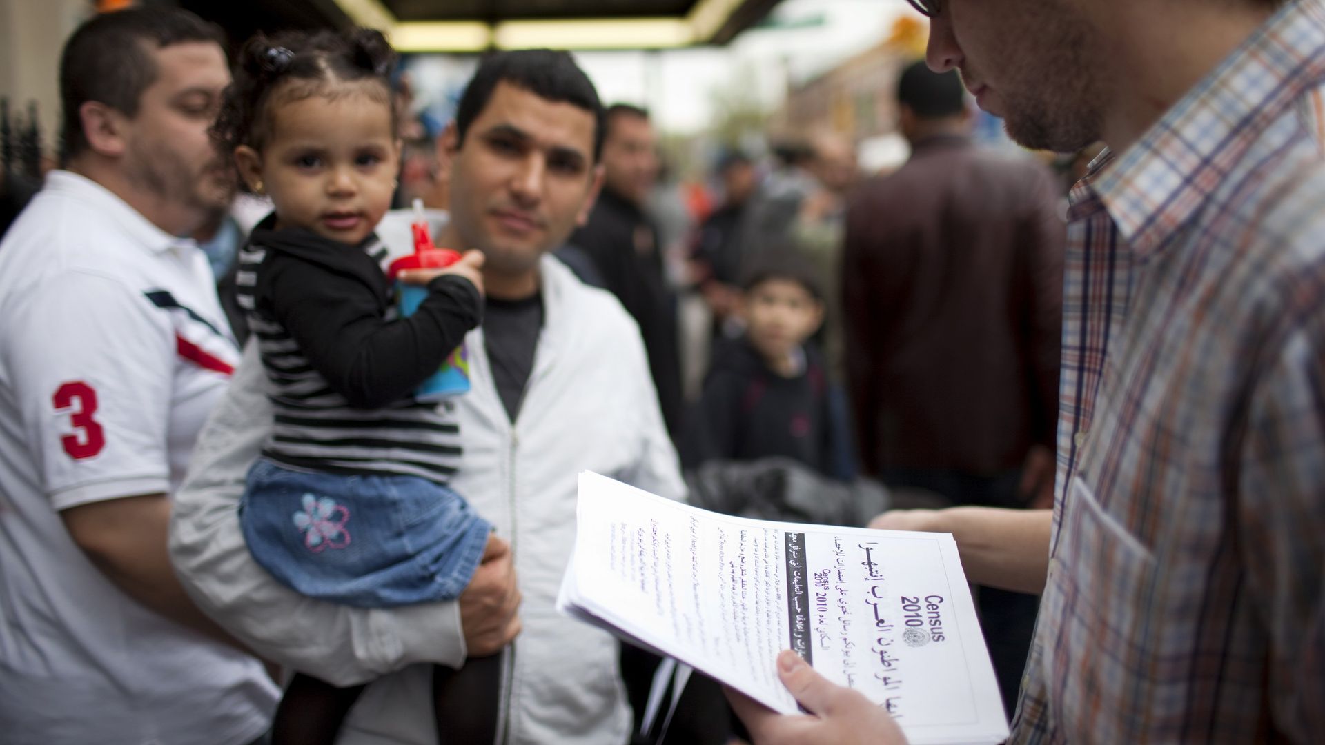 In this image, a man holds a toddler while another man hands out census papers.